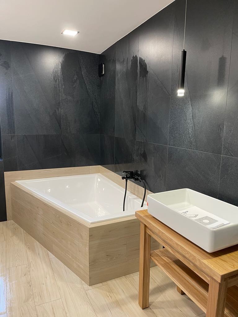 Nowoczesna łazienka z wanną obudowaną panelami imitującymi drewno, czarną armaturą i umywalką nablatową na drewnianym stoliku, ściany wyłożone ciemnoszarą płytką imitującą kamień.