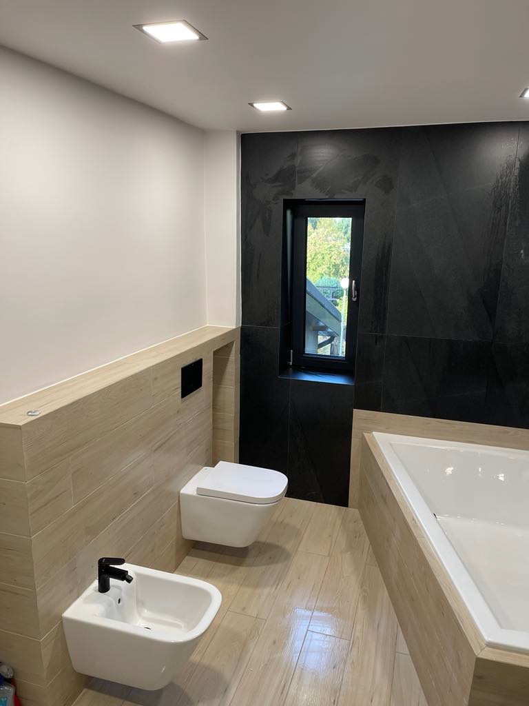 Nowoczesna łazienka z czarną ścianą z płytek imitujących łupek, drewnopodobnymi płytkami na podłodze i ścianach, wiszącą toaletą i bidetem z czarną armaturą oraz oknem z widokiem na zieleń.