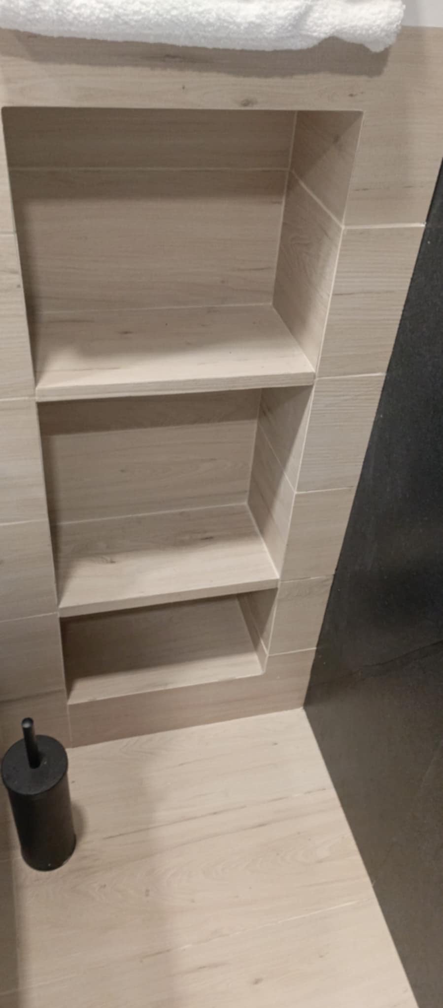 Półki wnękowe wyłożone płytkami imitującymi drewno, z czarnym akcentem ściany i toaletową szczotką w łazience po remoncie.