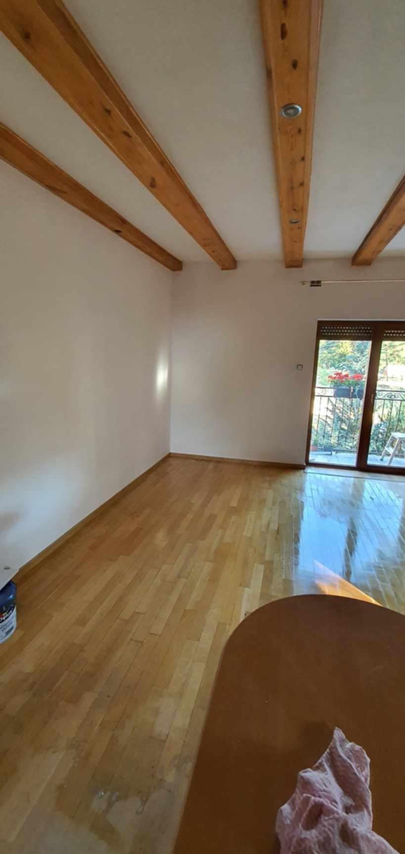 Pomalowane na jasny kolor ściany pokoju z drewnianymi belkami stropowymi, parkietem i wyjściem na balkon.