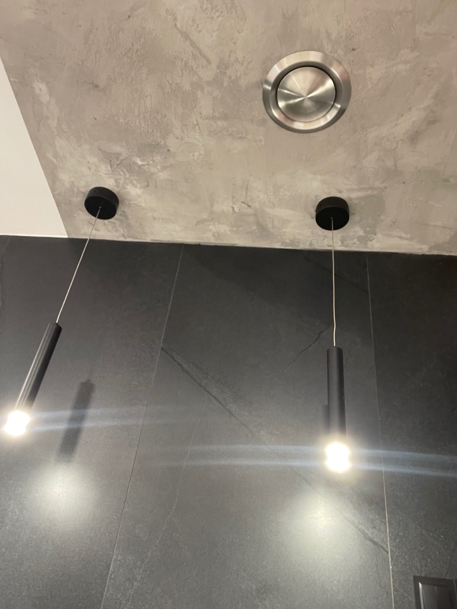 Szara ściana wykończona płytkami imitującymi kamień, oświetlona dwoma wiszącymi lampami z czarnymi, cylindrycznymi kloszami i okrągłym wlotem wentylacyjnym w suficie z efektem betonu.