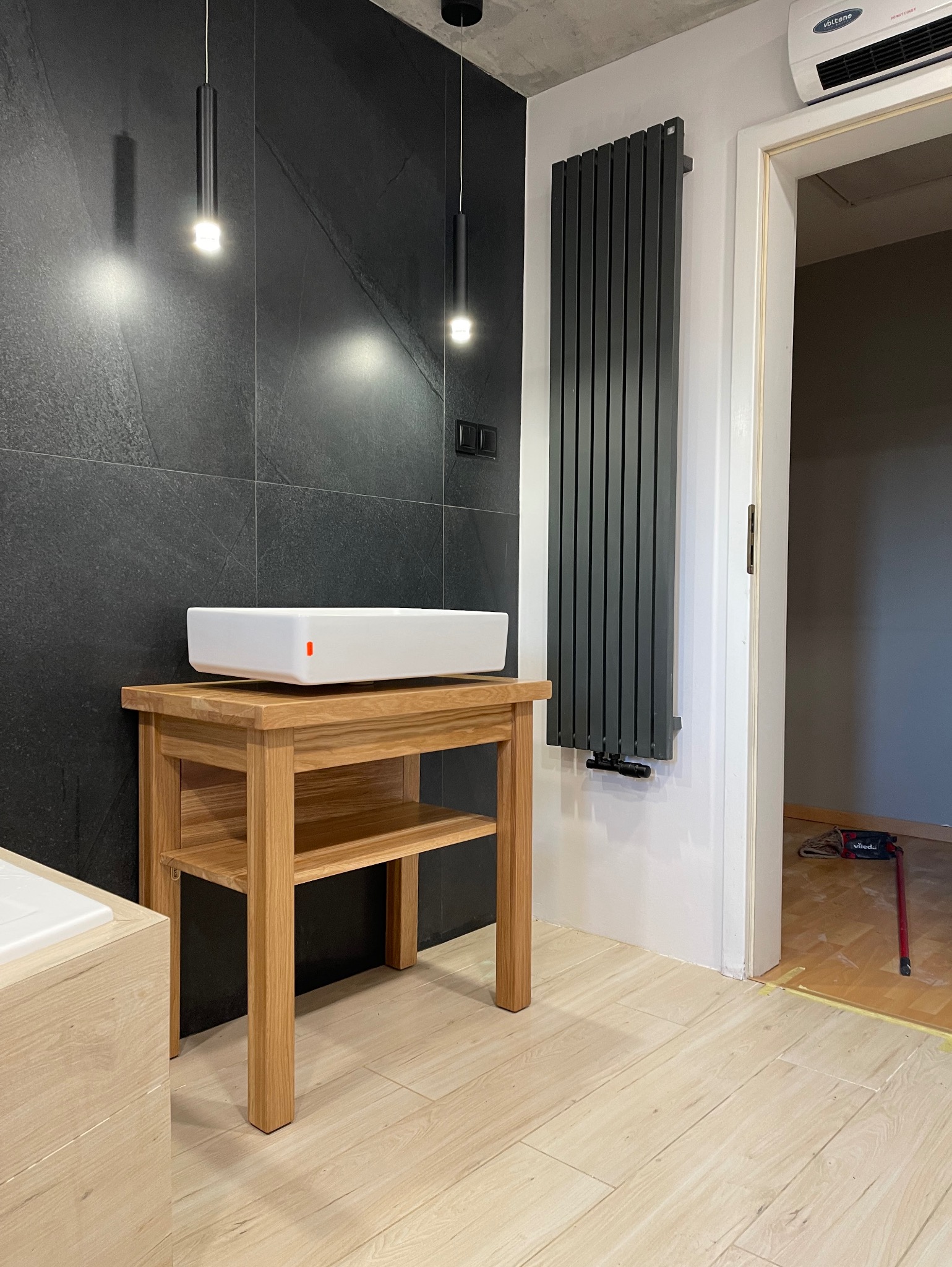 Nowoczesna łazienka z umywalką nablatową na drewnianym stoliku, czarne płytki na ścianie, grzejnik panelowy i podłoga wyłożona jasnymi panelami drewnopodobnymi.