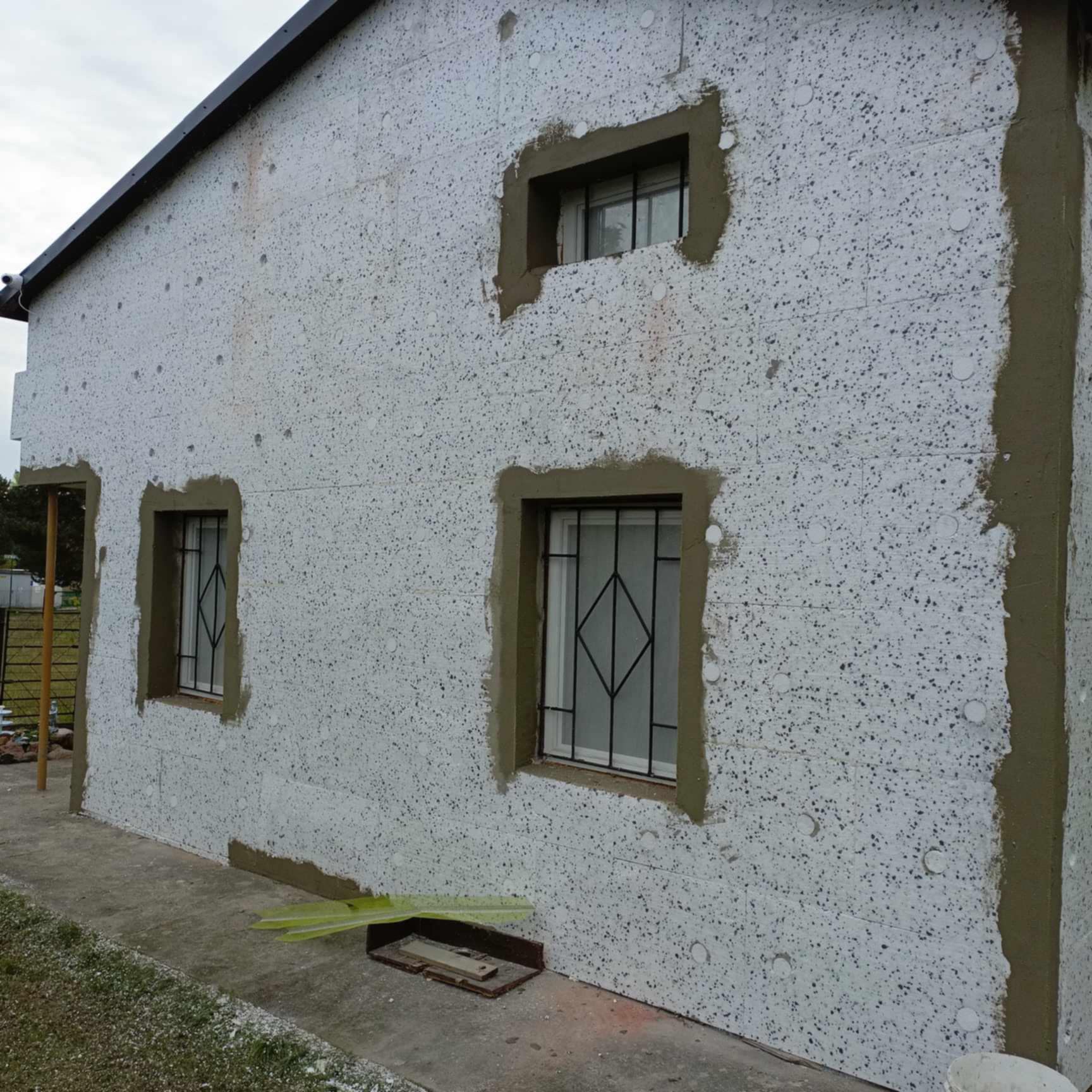 Ocieplenie elewacji budynku z widoczną warstwą styropianu i częściowo nałożoną zaprawą klejową wokół okien i na narożnikach, przygotowane do dalszych prac wykończeniowych.