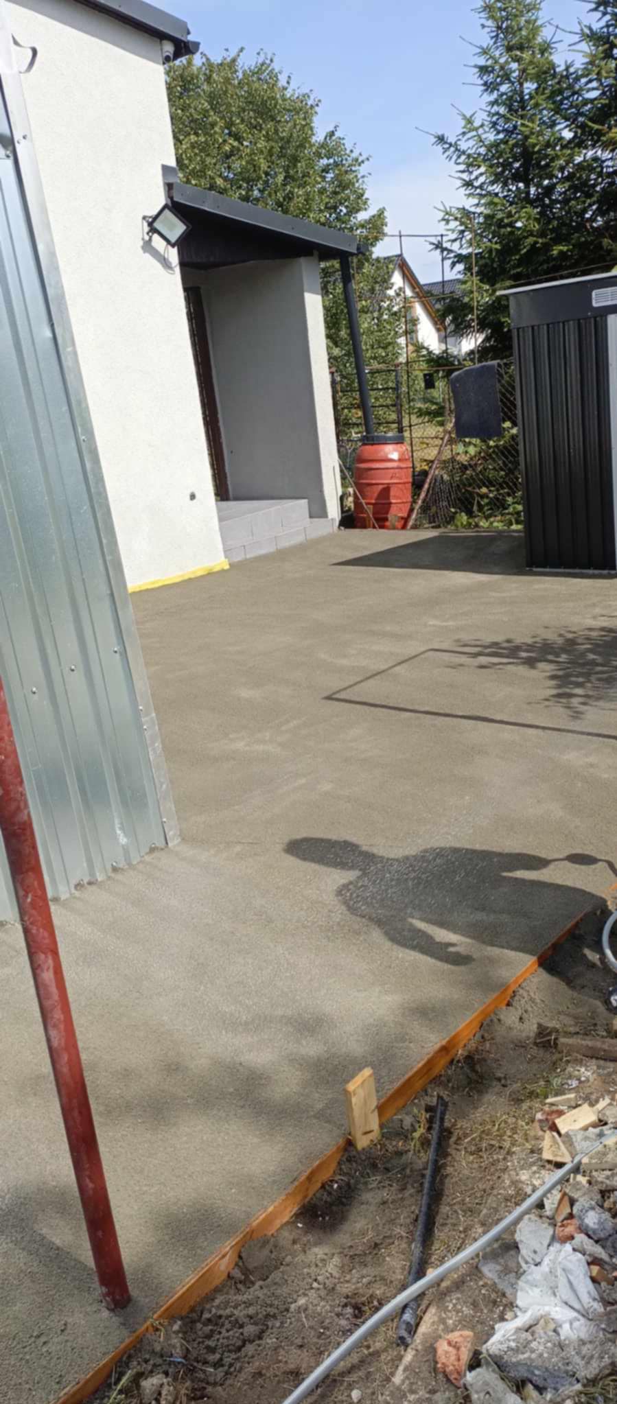 Świeżo wylana, szara betonowa wylewka tarasu przed domem, widoczne drewniane szalunki, cień osoby i częściowo metalowa osłona. W tle budynek z białym tynkiem i czerwona beczka.