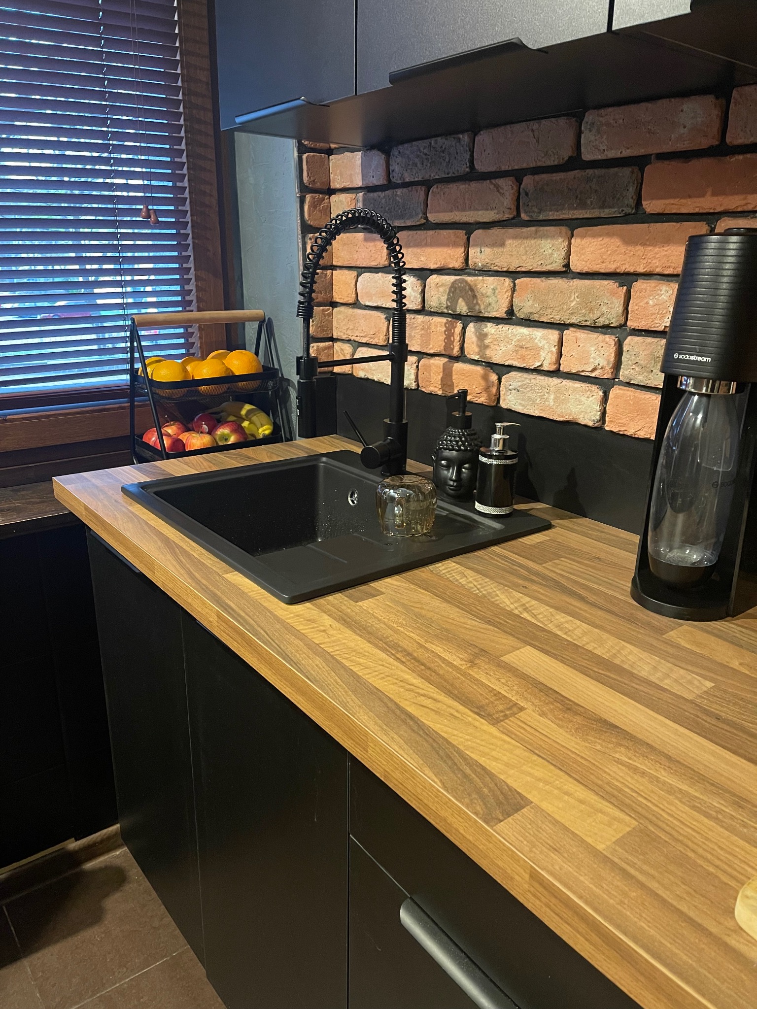 Nowoczesna kuchnia z czarnym zlewem, baterią w stylu retro i blatem z drewna, uzupełniona ceglaną ścianą i akcesoriami, takimi jak dozownik do mydła w kształcie głowy Buddy i saturator do wody.