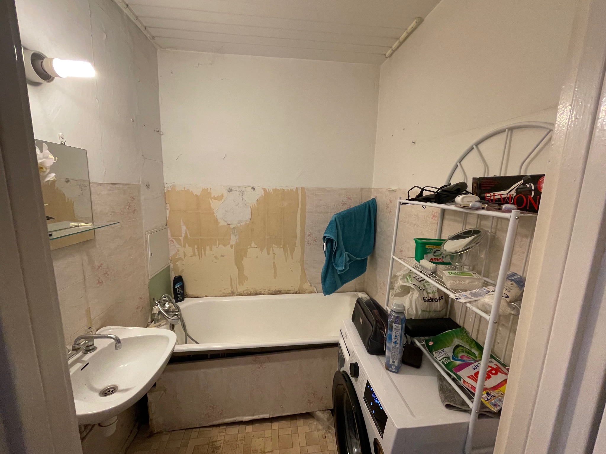 Łazienka przed remontem: widoczne stare płytki, ślady po usuniętych elementach, stara umywalka i wanna. Na wannie pralka i półka z detergentami. Widok na remontowaną łazienkę.