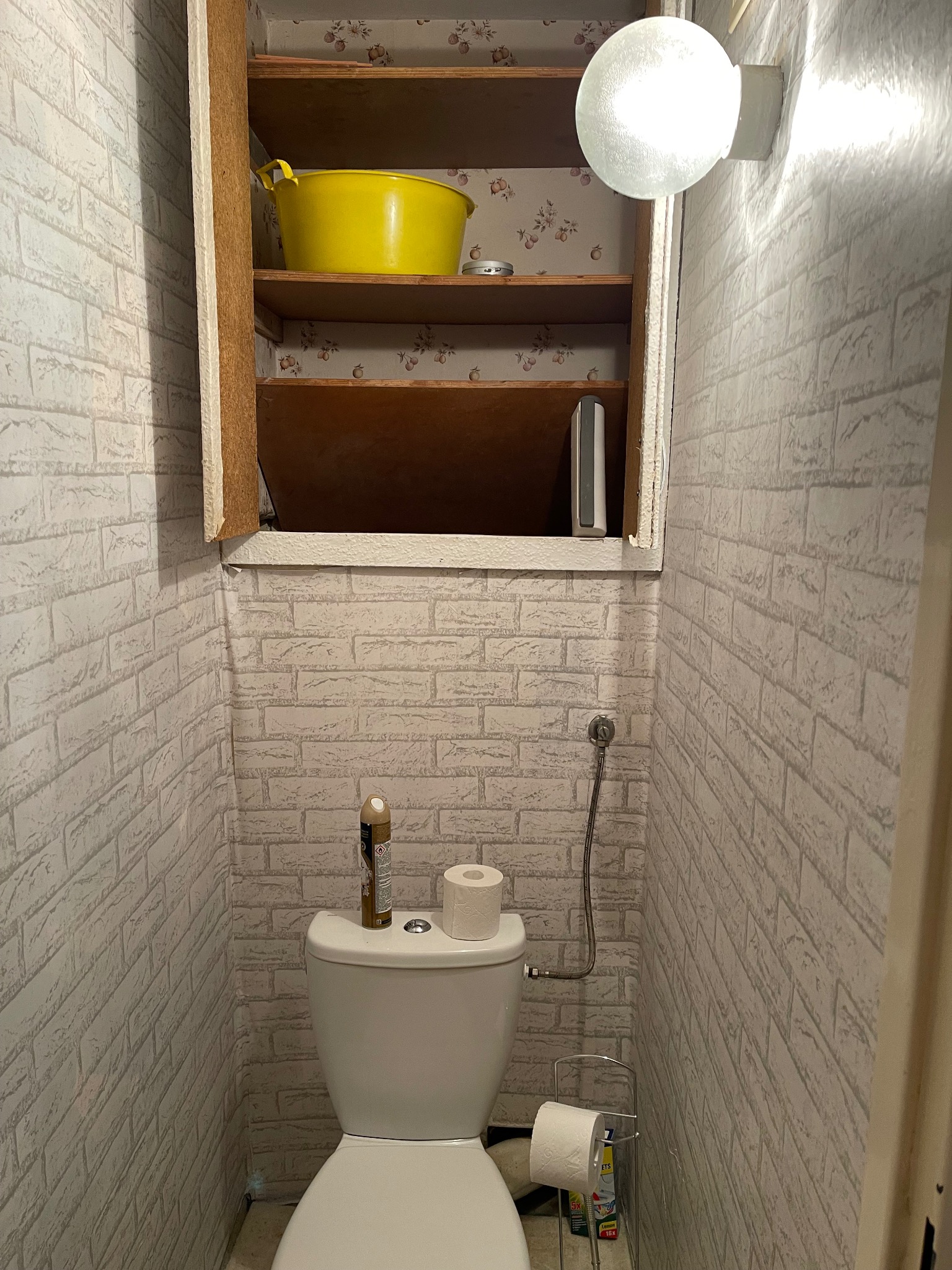 Wnętrze małej toalety z tapetą imitującą cegłę, otwartą szafką z żółtą miską i półkami z wzorzystą tapetą w tle. Nad szafką okrągła lampa.