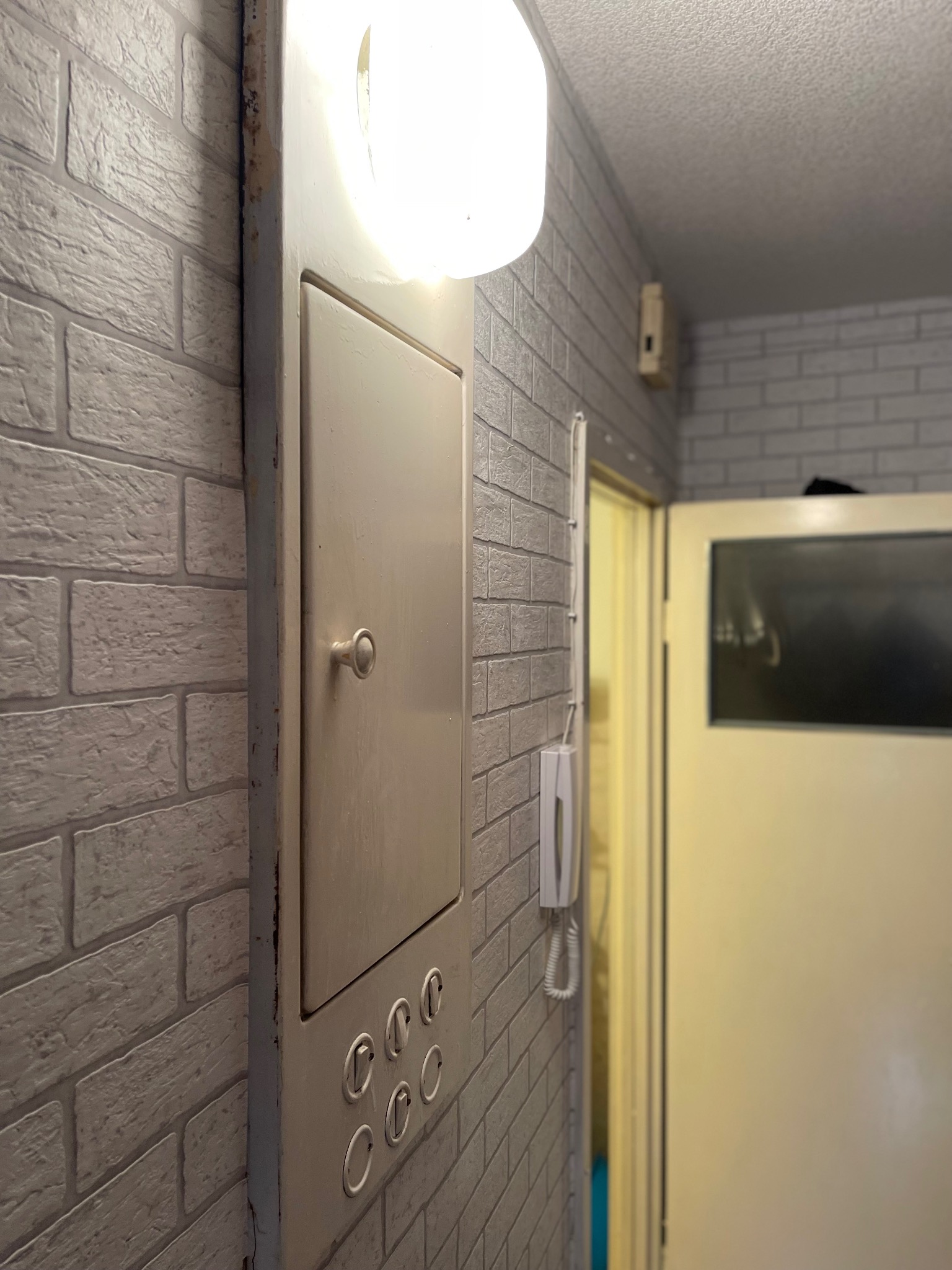 Szara ściana z cegły z metalową skrzynką z oświetleniem i przyciskami. W tle widoczne drzwi i telefon na ścianie. Ujęcie z bliska, z perspektywy lekko od dołu.