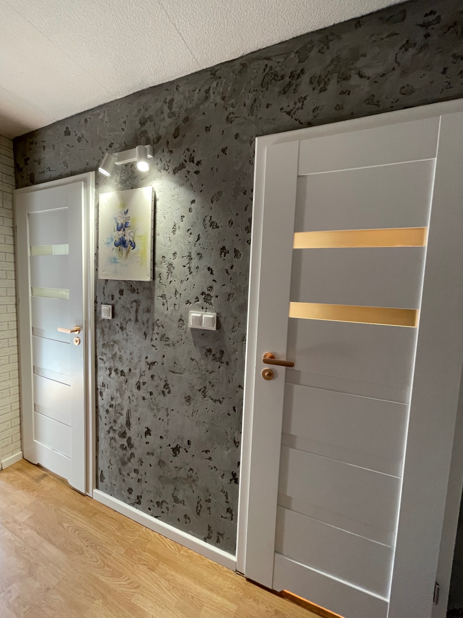 Dwa białe, nowoczesne drzwi z przeszkleniami na tle ściany z dekoracyjnym tynkiem w odcieniach szarości. Na ścianie obraz i włączniki światła. Podłoga drewniana.