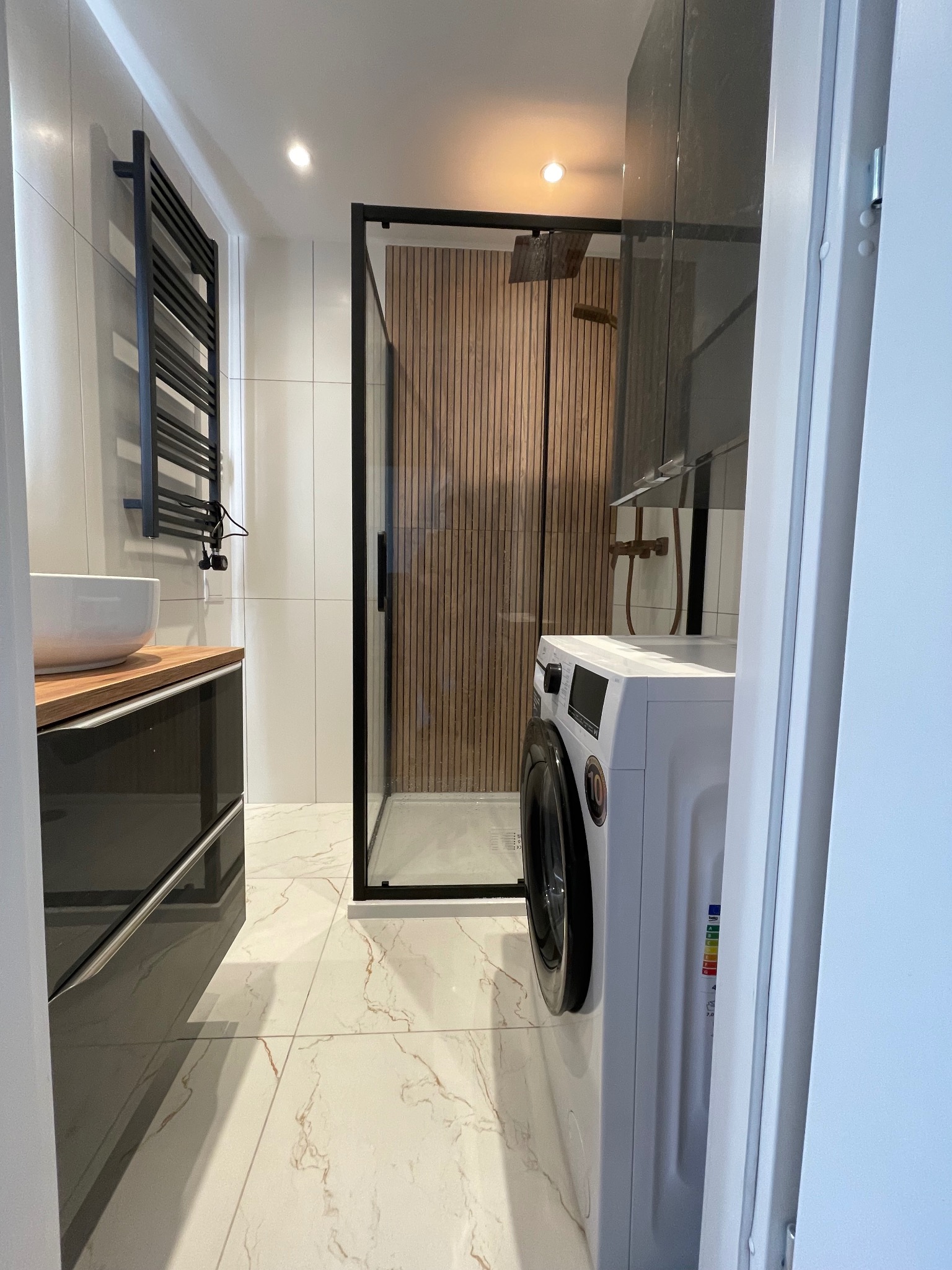 Nowoczesna łazienka z kabiną prysznicową typu walk-in z drewnianym panelem, umywalką nablatową i czarnym grzejnikiem drabinkowym. Marmurowe płytki podłogowe.