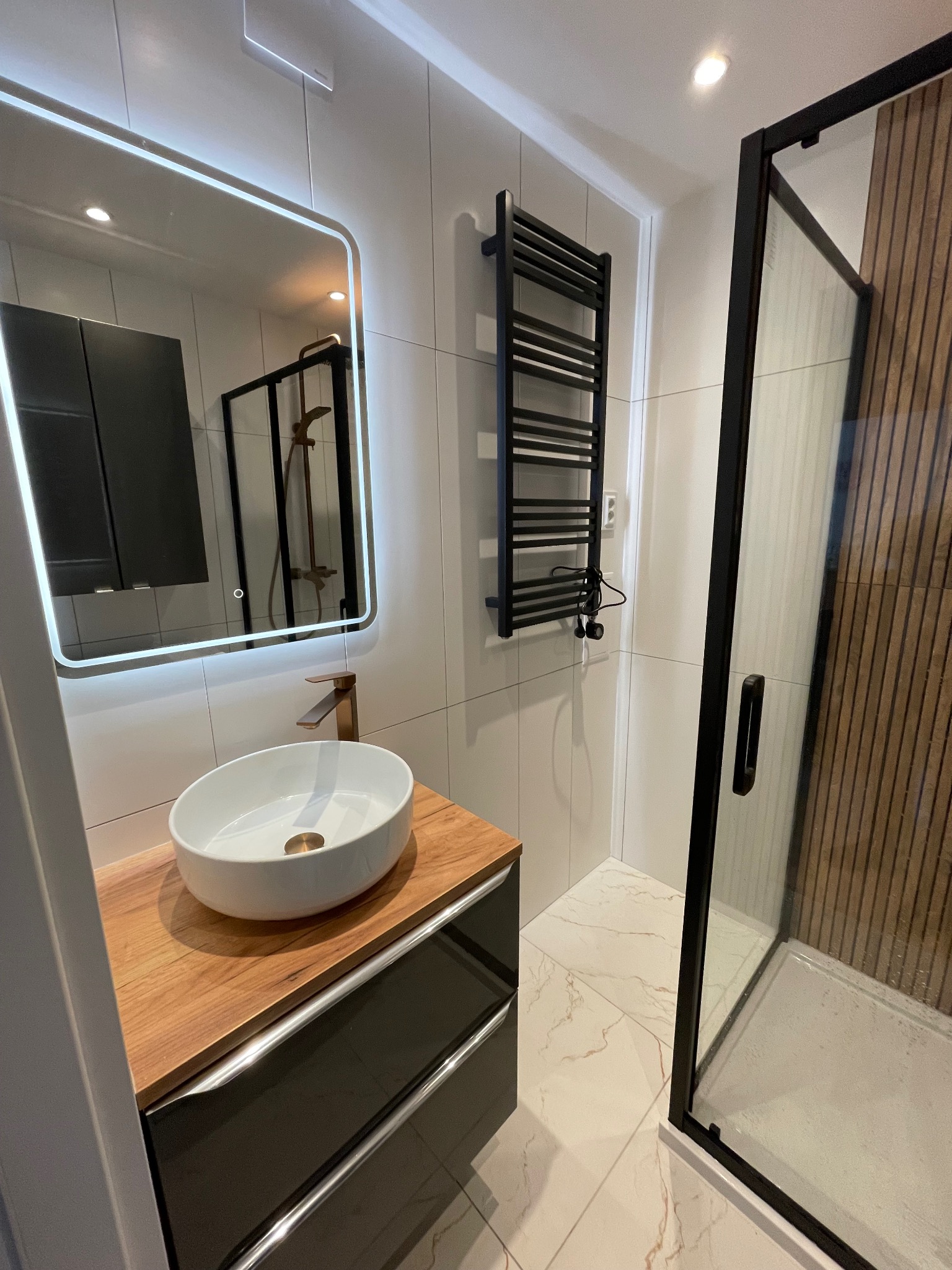 Nowoczesna łazienka po remoncie: białe płytki, umywalka nablatowa na drewnianym blacie, czarna kabina prysznicowa i grzejnik drabinkowy. Lustro z podświetleniem LED.