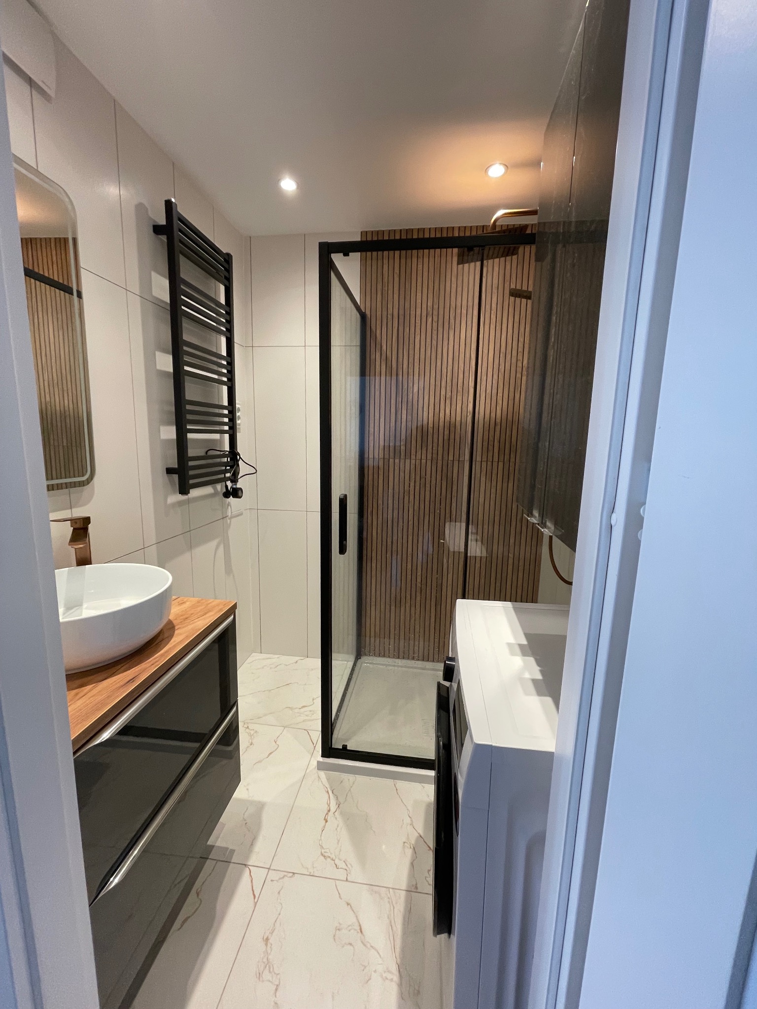 Nowoczesna łazienka z kabiną prysznicową typu walk-in, umywalką nablatową na drewnianym blacie i grzejnikiem drabinkowym w czarnym kolorze.