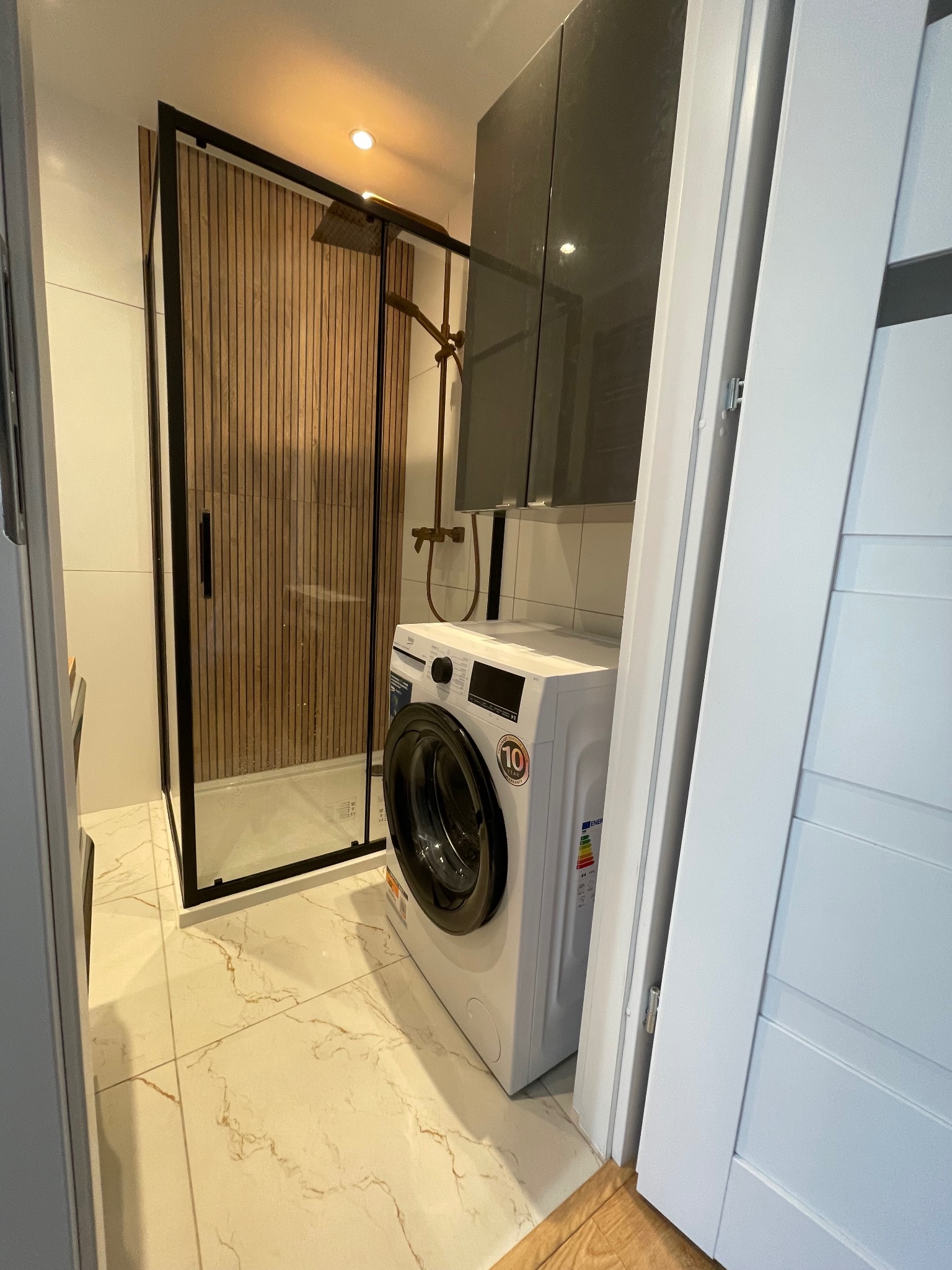 Nowoczesna łazienka z kabiną prysznicową typu walk-in z drewnianymi panelami, pralką i szafkami w szarym odcieniu. Marmurowe płytki podłogowe.