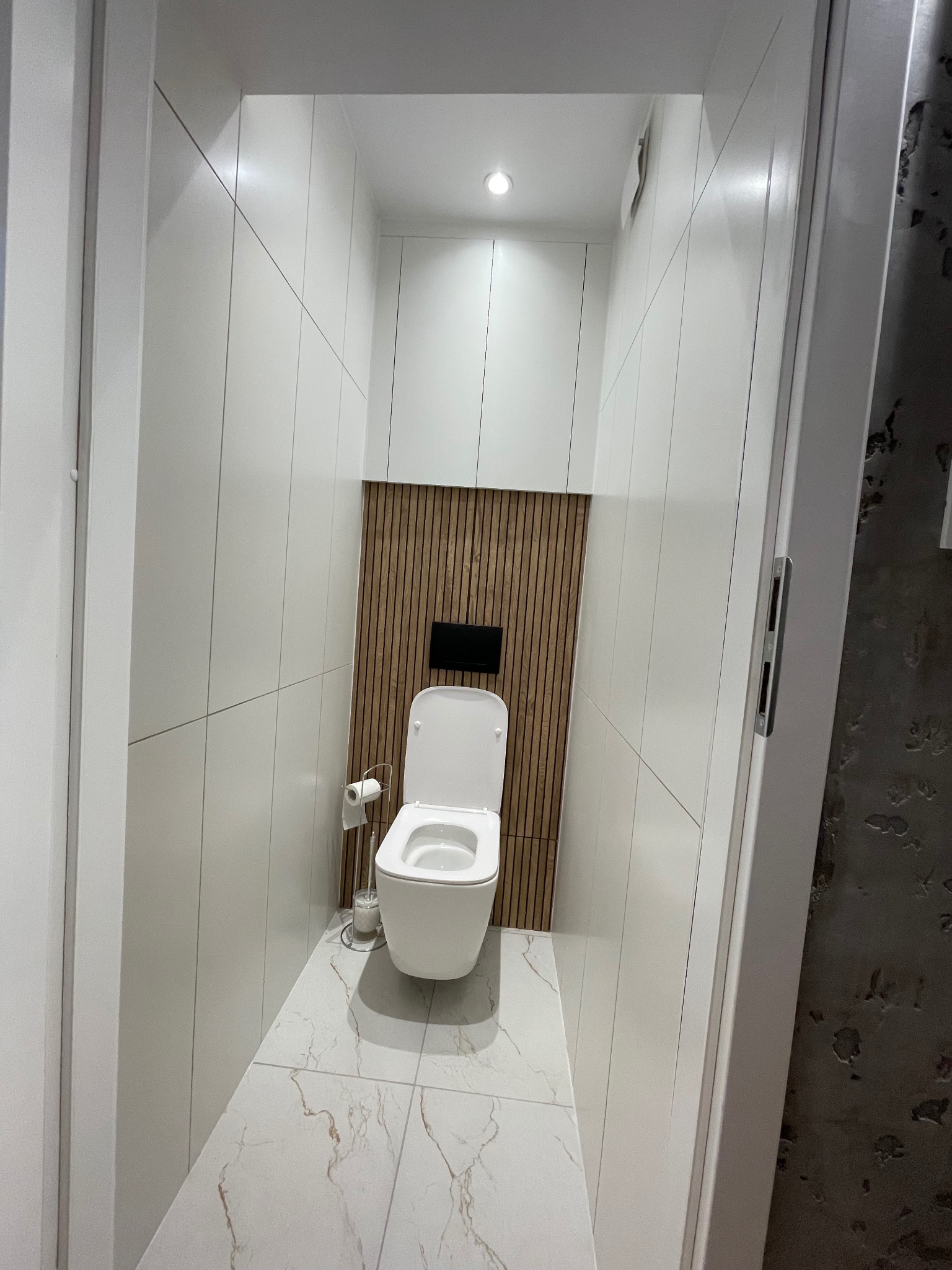 Nowoczesna toaleta z białymi panelami ściennymi i drewnianą ścianą akcentującą. Marmurowe płytki podłogowe i minimalistyczny design.
