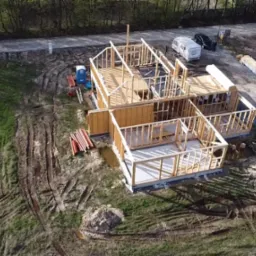 Budowa domu w technologii szkieletowej z widocznymi elementami konstrukcyjnymi ścian i dachu, z perspektywy lotniczej, na działce z widocznym traktorem, przyczepą kempingową i przenośną toaletą.