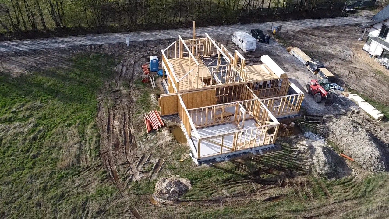 Budowa domu w technologii szkieletowej z widocznymi elementami konstrukcyjnymi ścian i dachu, z perspektywy lotniczej, na działce z widocznym traktorem, przyczepą kempingową i przenośną toaletą.