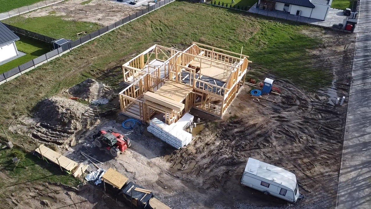 Wznoszenie konstrukcji szkieletowej domu z widokiem z góry, widoczny czerwony podnośnik teleskopowy, przyczepa kempingowa i tymczasowa toaleta na placu budowy.