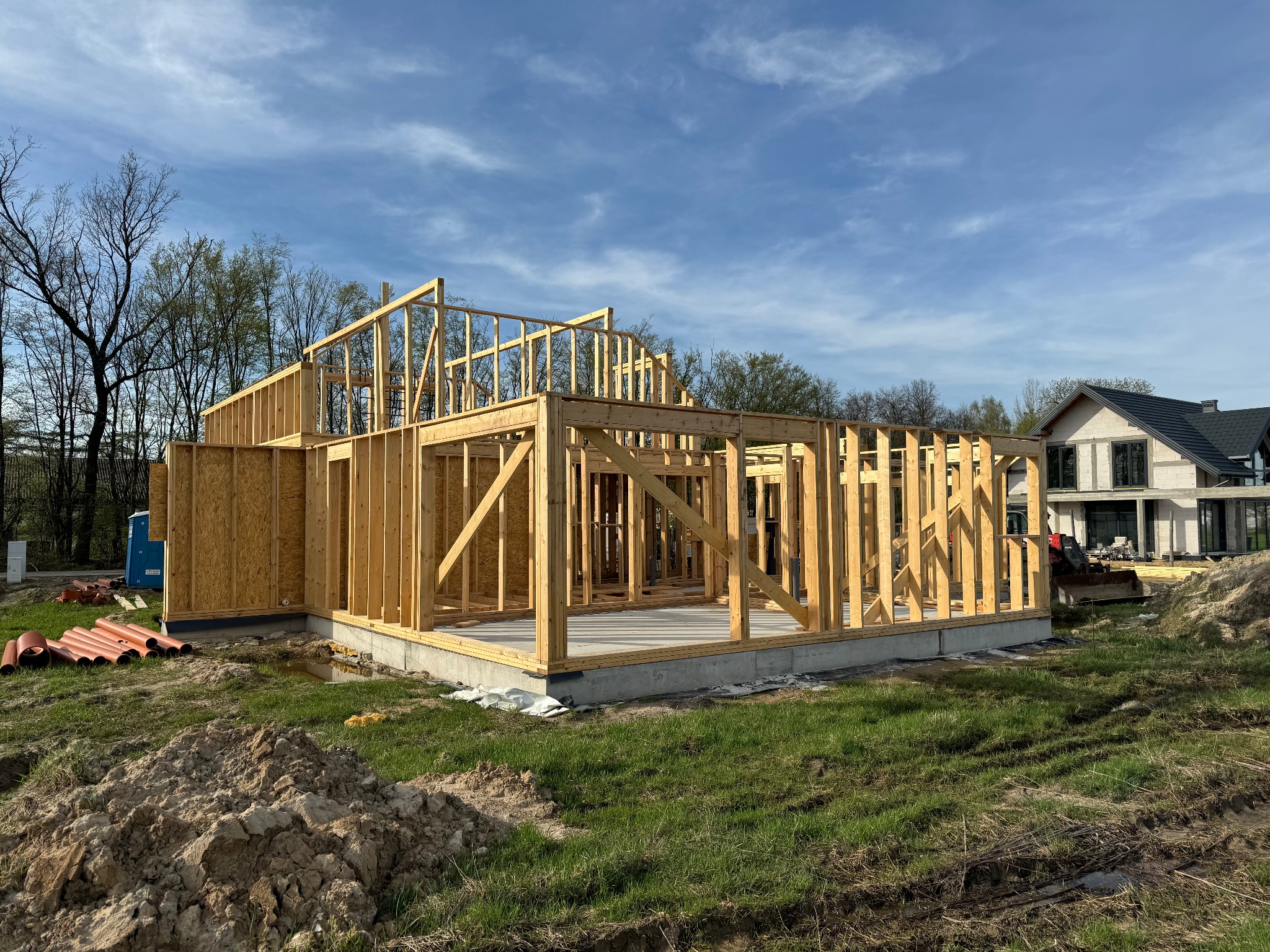 Surowy szkielet domu w trakcie budowy, widoczna betonowa podstawa, drewniane ramy ścian i dachu, na zewnątrz składowane rury i widoczny inny budynek w tle.
