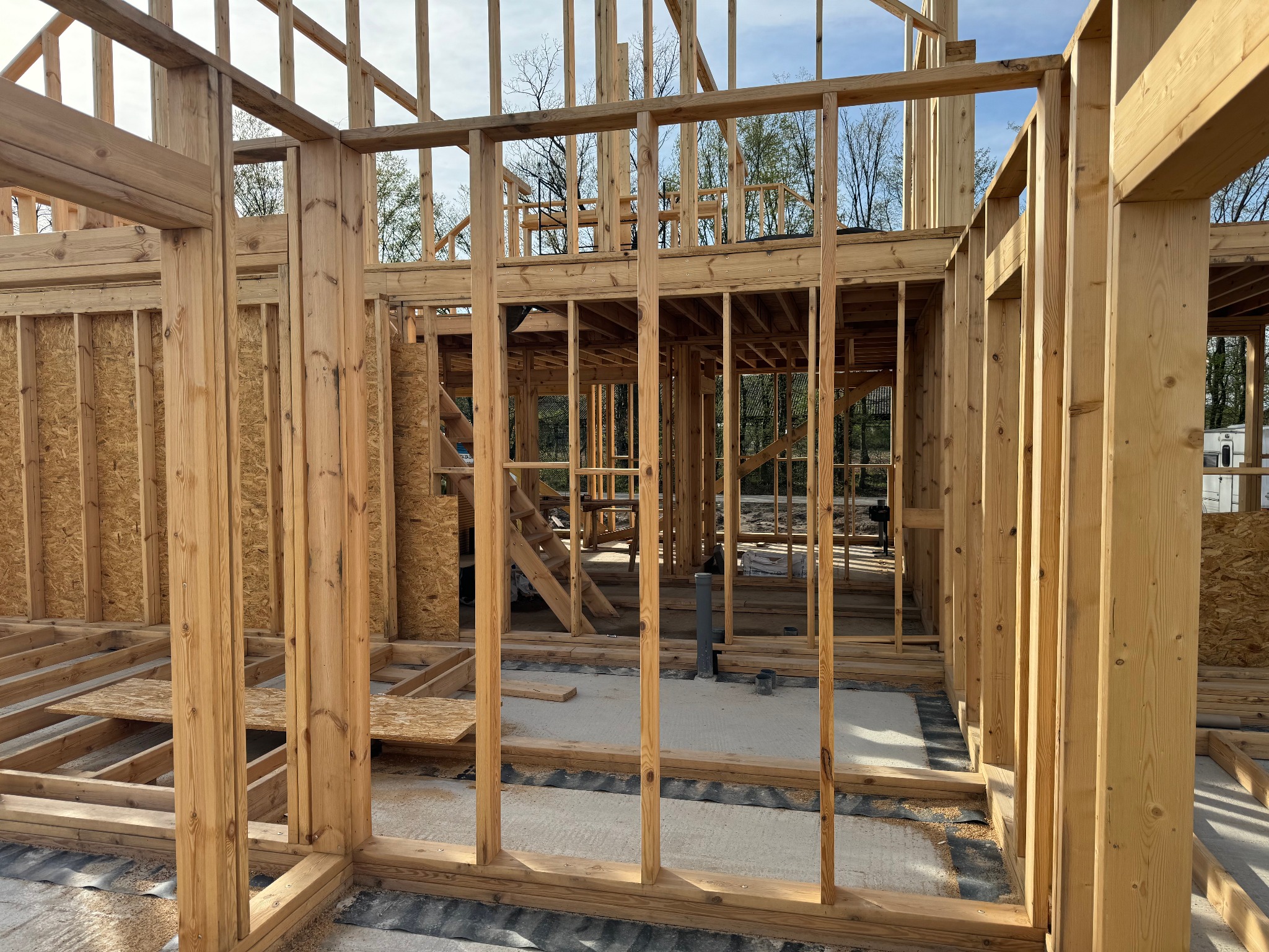 Surowa konstrukcja drewniana domu w trakcie budowy, widoczne słupy, belki i podłogi, z fragmentami płyty OSB, na tle zieleni i błękitnego nieba.
