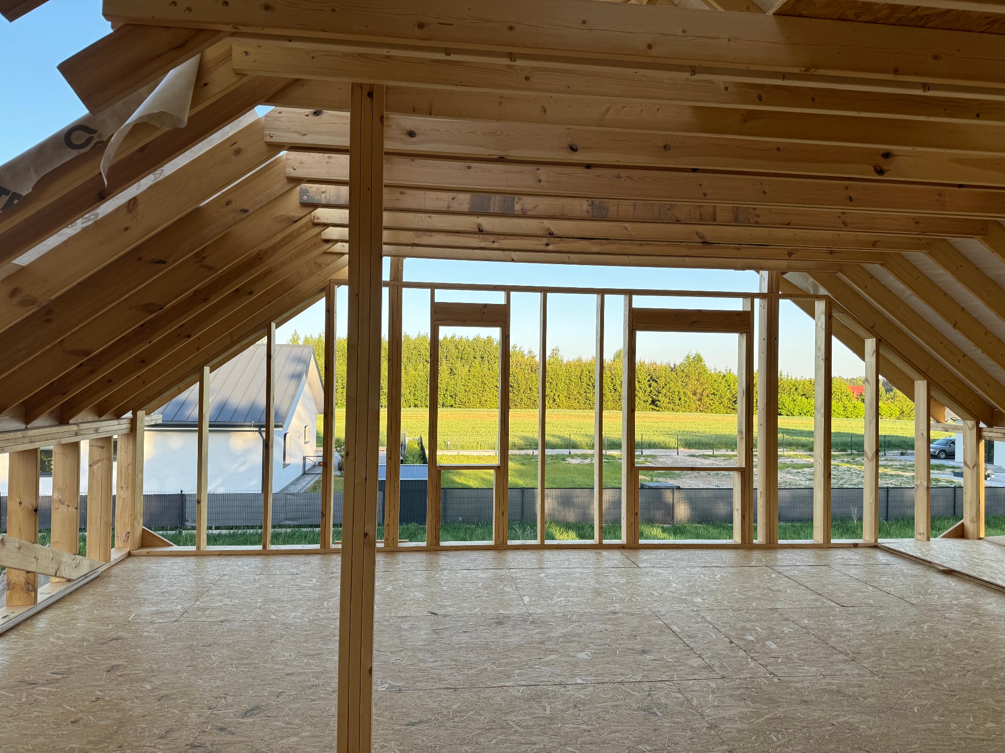 Wnętrze w trakcie budowy w Wołyńce-Kolonia: widok na szkielet konstrukcji drewnianej domu, z podłogą z płyt OSB i widokiem na zewnątrz przez otwory okienne.