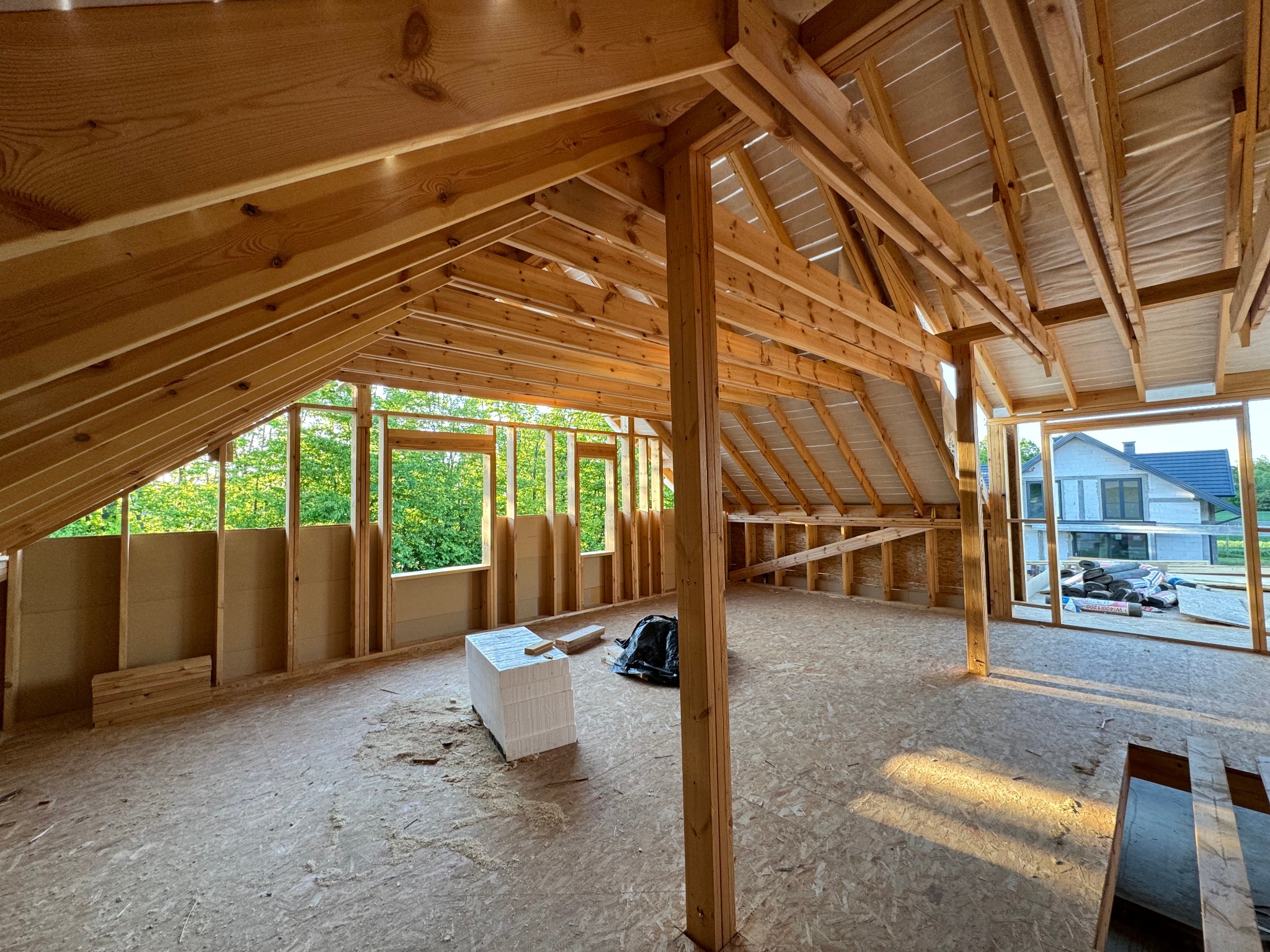 Wnętrze w trakcie budowy z drewnianą konstrukcją dachu, widoczne okna z zielenią na zewnątrz i materiały budowlane na podłodze.