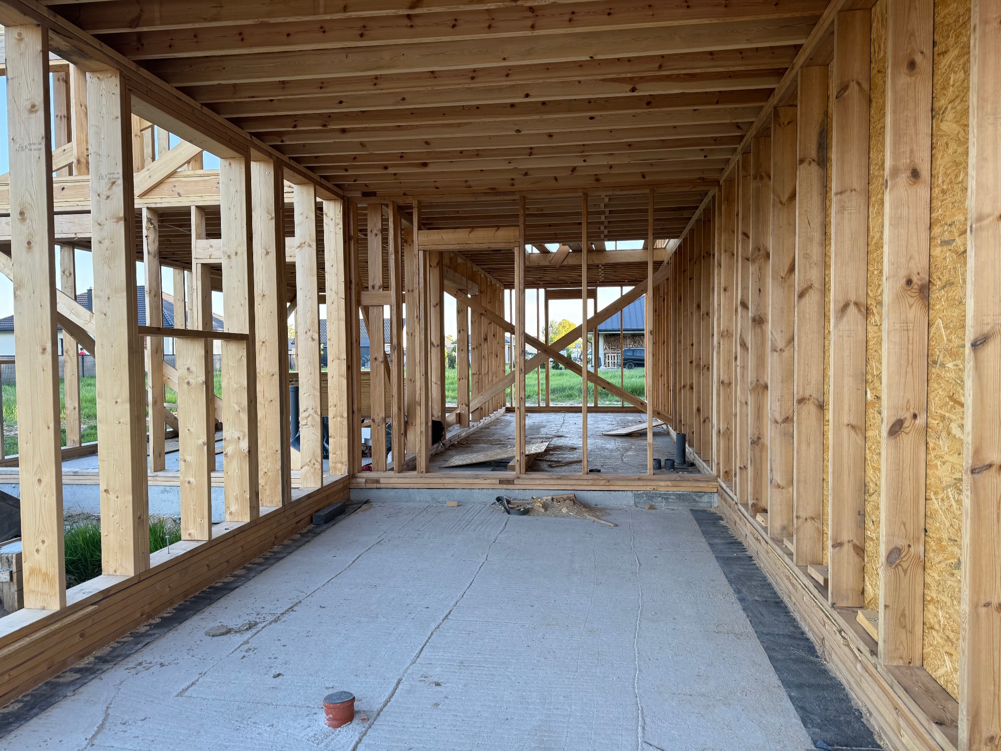 Wnętrze surowego domu szkieletowego w trakcie budowy, widoczne drewniane ramy ścian i stropu, betonowa podłoga z widocznymi śladami po narzędziach.