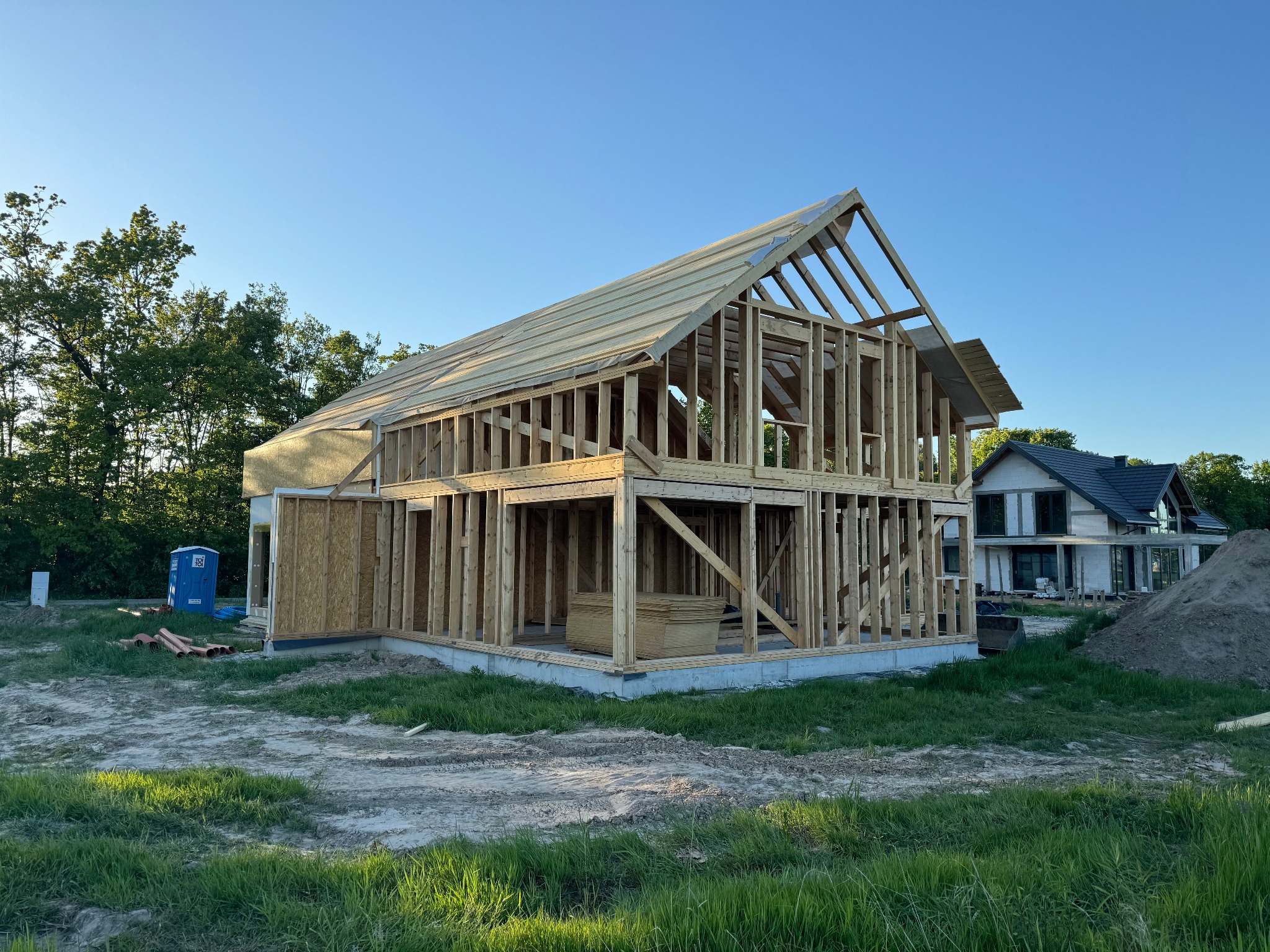 Wznoszenie konstrukcji domu z drewnianych belek na fundamencie betonowym, widoczne składowane płyty OSB, przenośna toaleta i sąsiedni dom w tle.