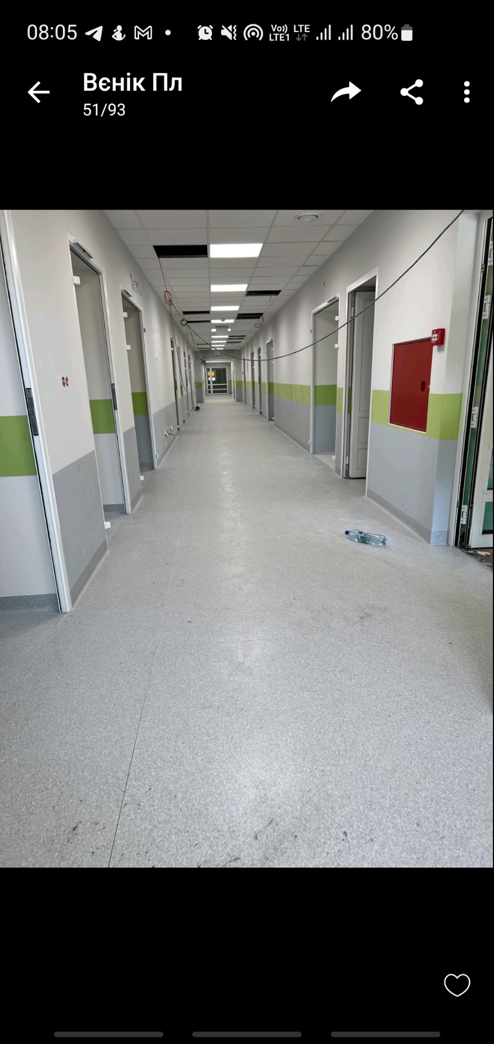 Długi korytarz w budynku publicznym z szarą wykładziną podłogową, białymi ścianami z zielonymi akcentami, czerwona szafka hydrantowa na ścianie i butelka wody na podłodze.