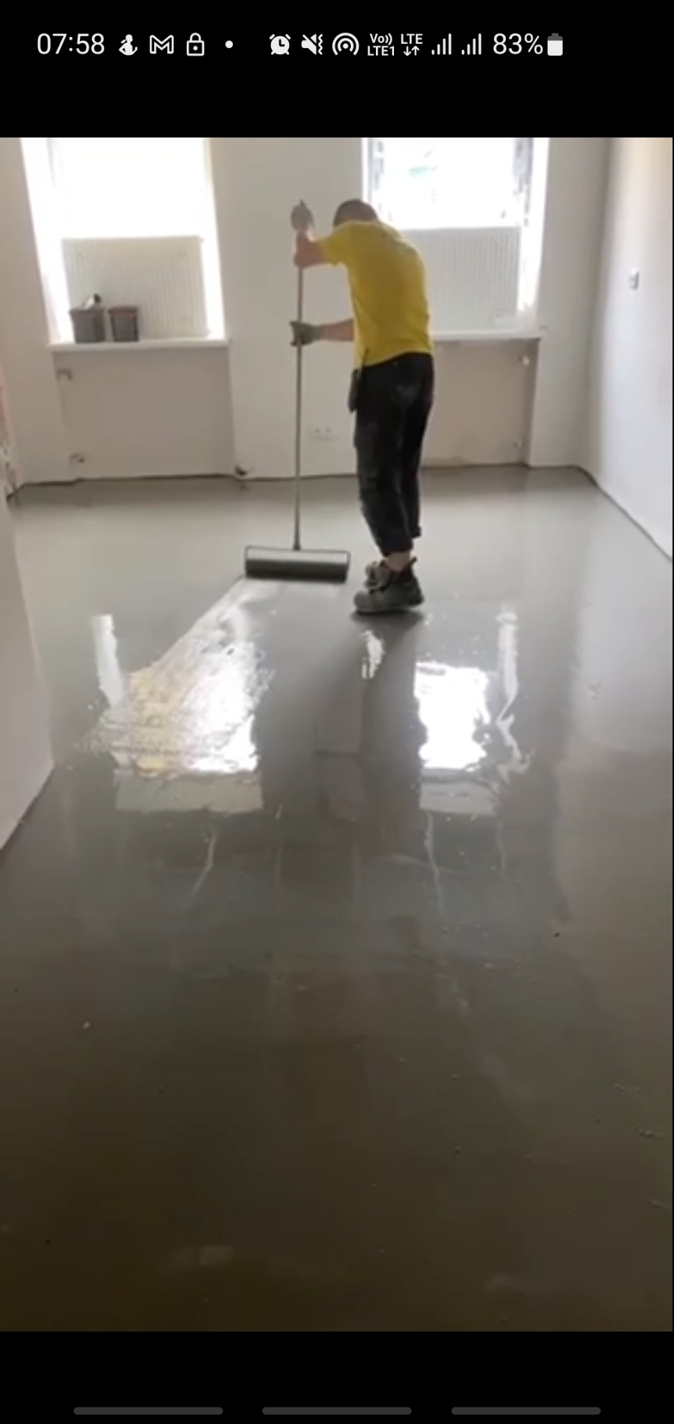 Pracownik w żółtej koszulce rozprowadza szpachlą samopoziomującą masę na podłodze w jasnym pomieszczeniu z grzejnikami pod oknami.