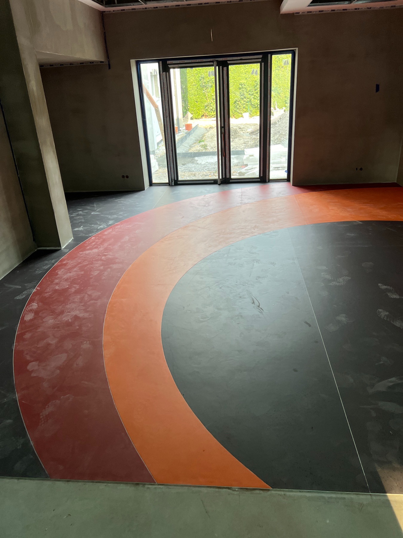 Wielobarwna wykładzina podłogowa z wzorem łuku w nowym budynku, widoczne ślady budowlane i okno z widokiem na zieleń.