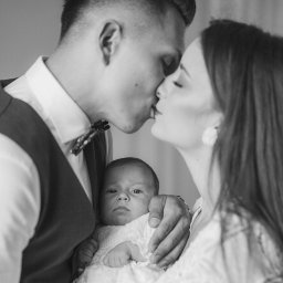 Klaudia Wiśniowska Media - Czarno-białe ujęcie całującej się pary trzymającej niemowlę w białej, koronkowej sukience, dziecko patrzy prosto w obiektyw.