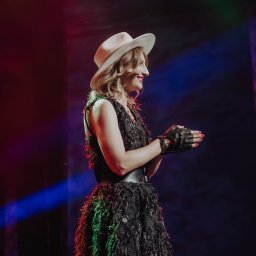 Klaudia Wiśniowska Media - Kobieta z profilu w czarnej sukience z frędzlami, beżowym kapeluszu i skórzanych rękawiczkach bez palców, uśmiechnięta, oświetlona kolorowymi światłami scenicznymi.