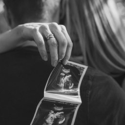 Klaudia Wiśniowska Media - Czarno-białe zdjęcie pary obejmującej się, trzymającej serię zdjęć USG w dłoni z widocznymi obrączkami.