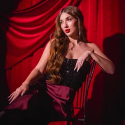 Portret kobiety w cekinowym gorsecie i burgundowej spódnicy, siedzącej na krześle na tle czerwonych drapowanych zasłon, z wyraźnym makijażem i falowanymi włosami.