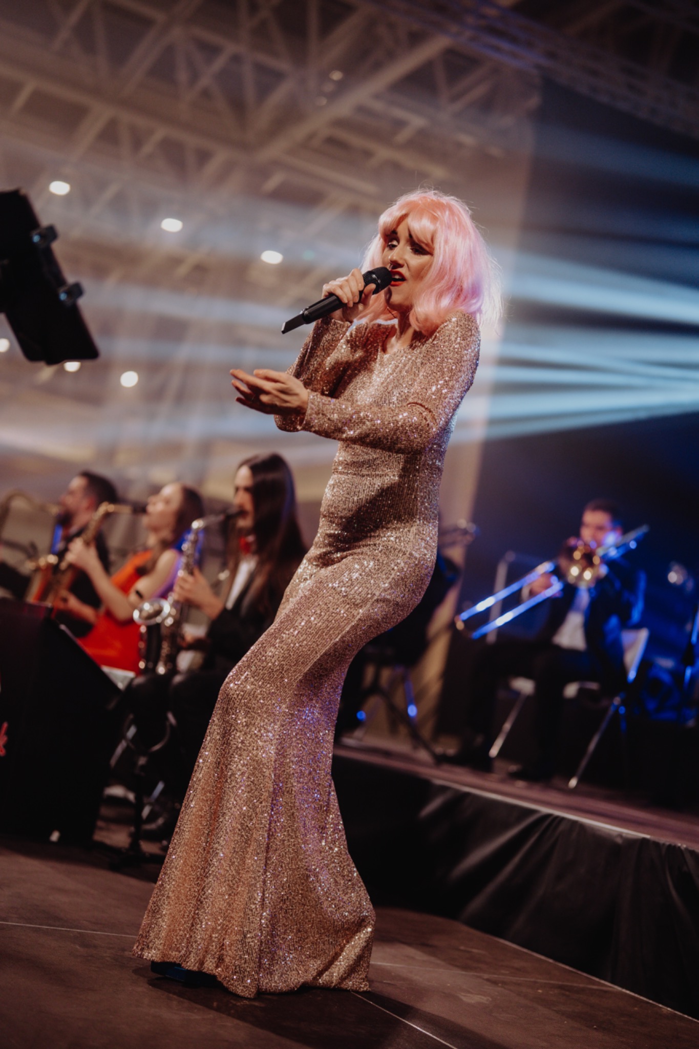 Wokalistka w błyszczącej sukni i różowej peruce śpiewa do mikrofonu na scenie z zespołem muzycznym w tle, oświetlona niebieskim światłem.