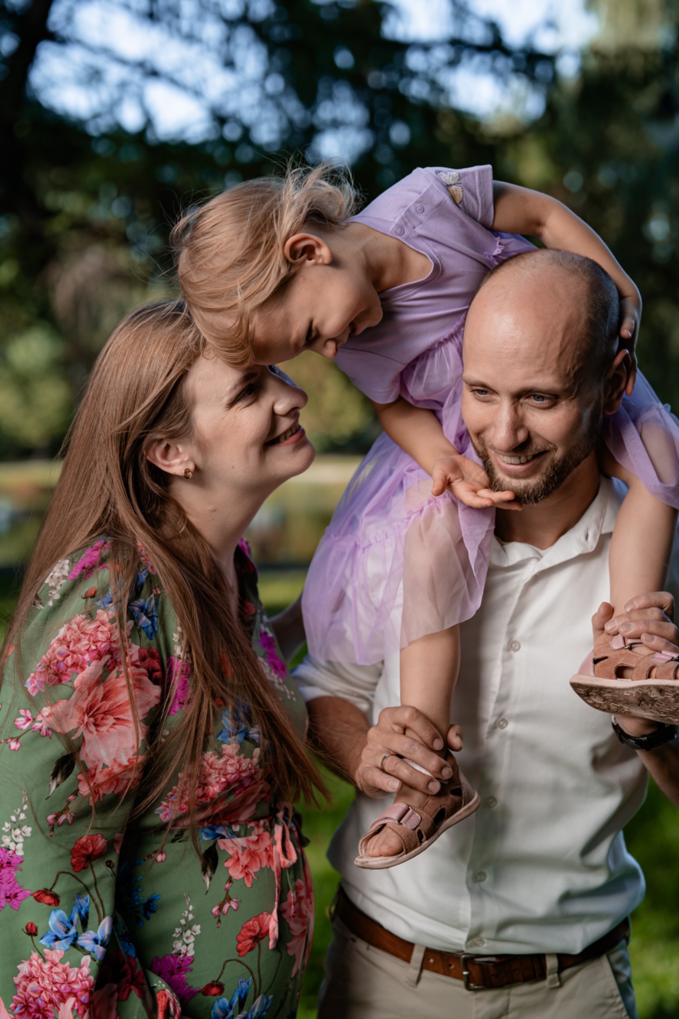 Uśmiechnięta rodzina w parku: córka na ramionach ojca, matka patrzy z czułością, dziewczynka w liliowej sukience, kobieta w kwiecistej sukience.