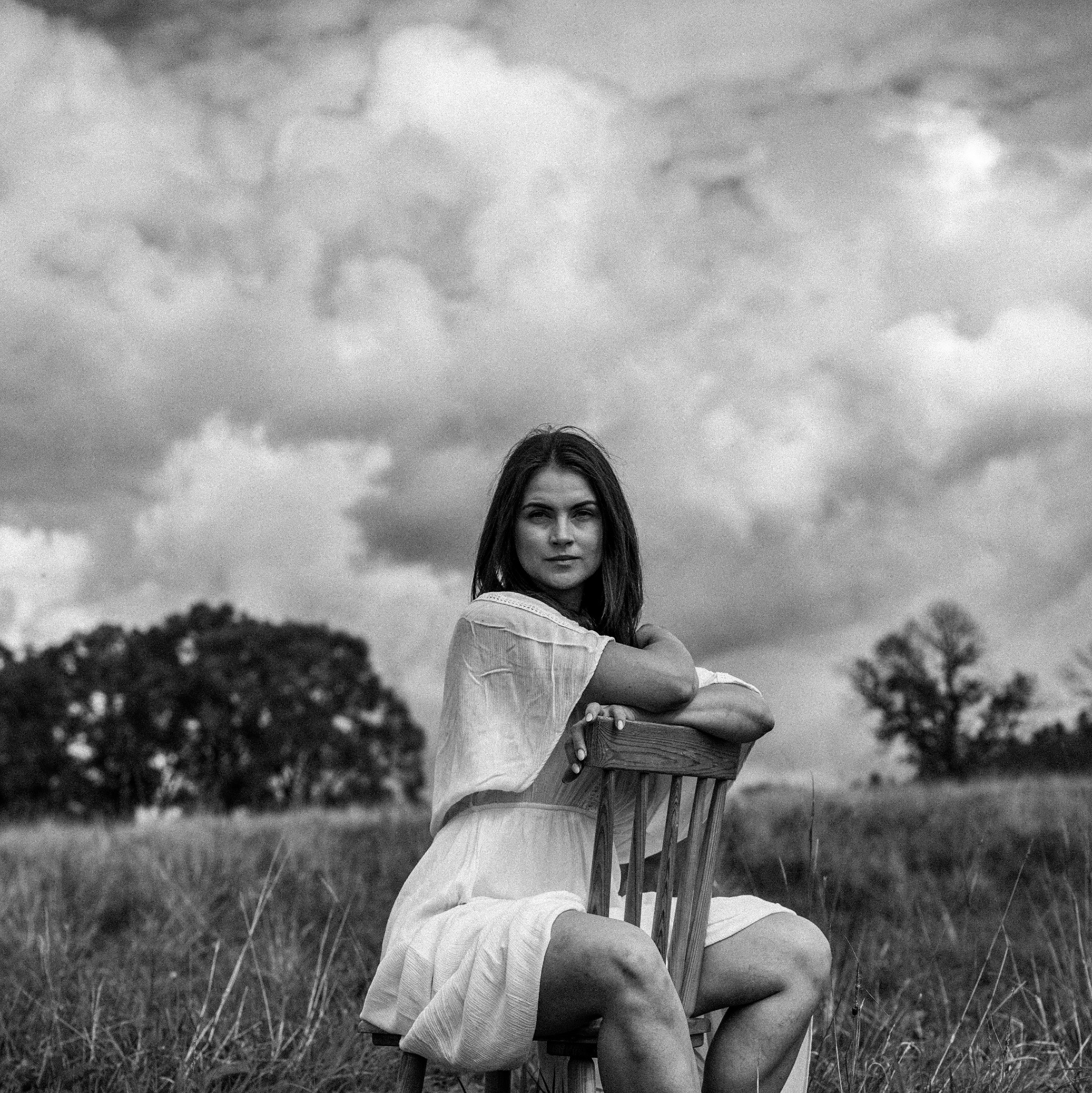 Czarno-białe zdjęcie kobiety w zwiewnej sukience siedzącej na drewnianym krześle na tle łąki i pochmurnego nieba.