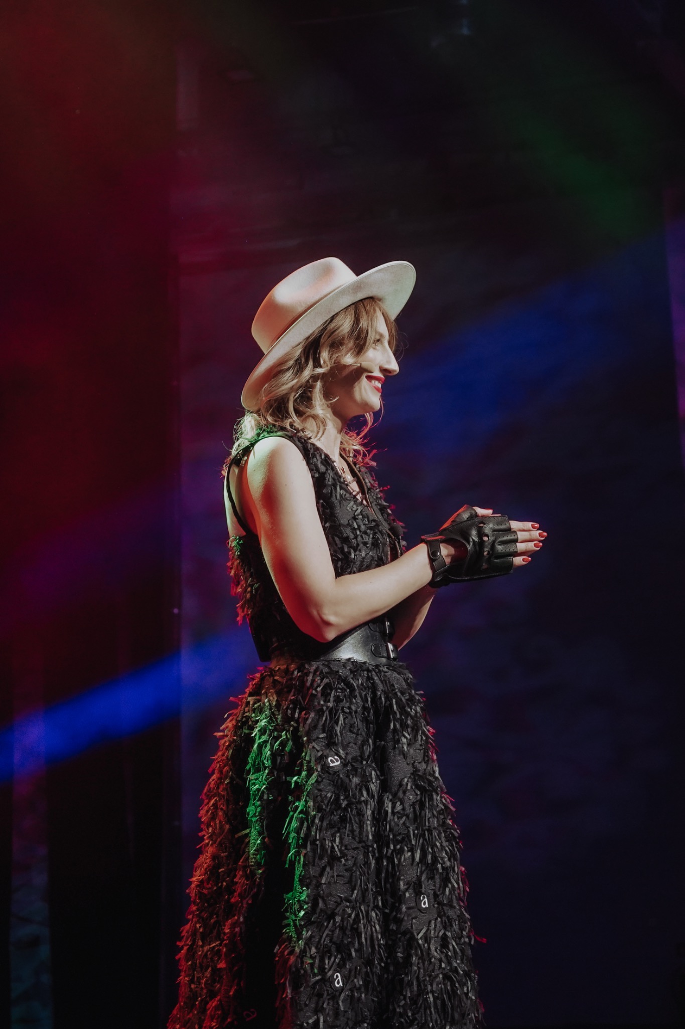 Kobieta z profilu w czarnej sukience z frędzlami, beżowym kapeluszu i skórzanych rękawiczkach bez palców, uśmiechnięta, oświetlona kolorowymi światłami scenicznymi.
