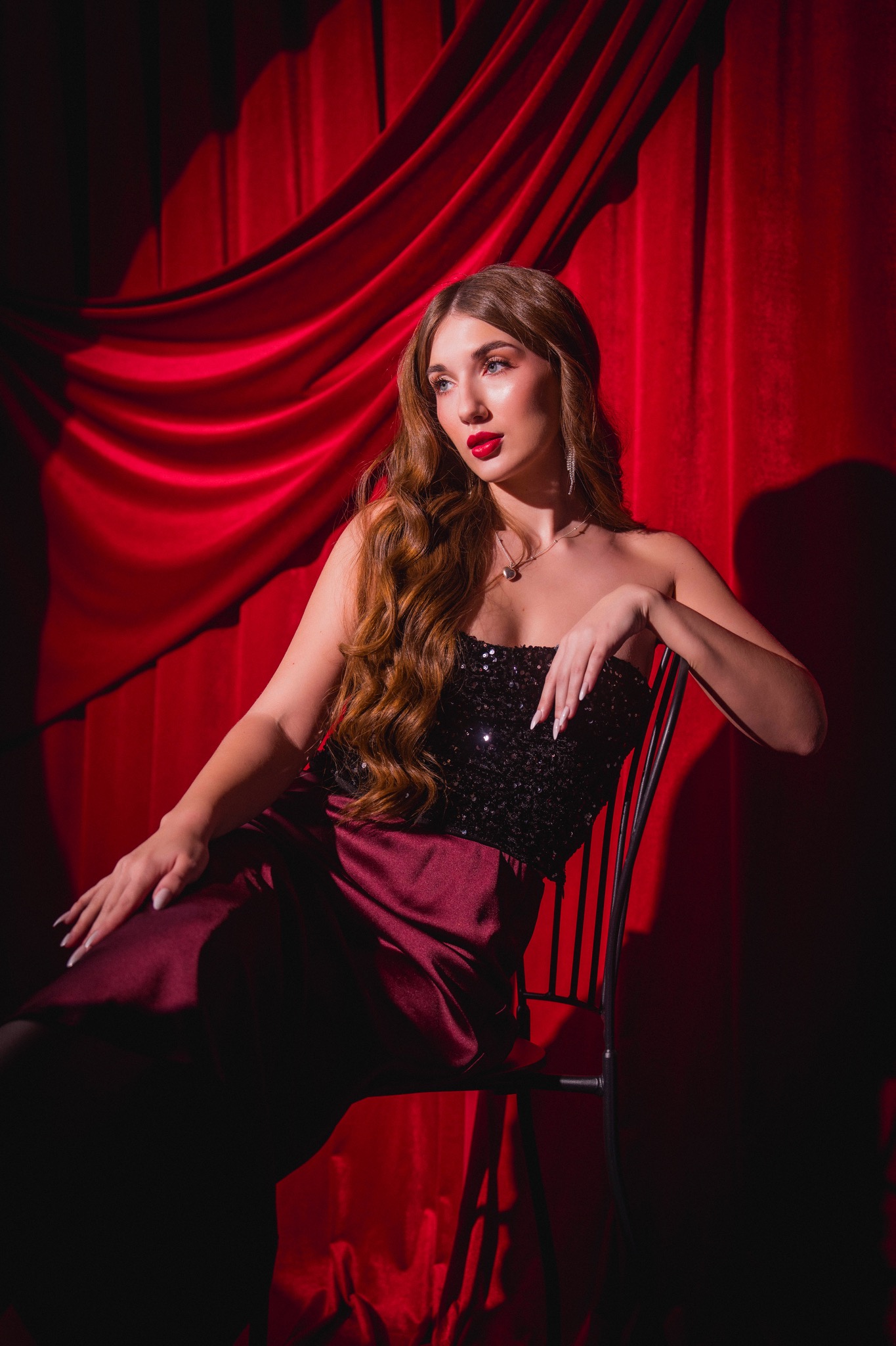 Portret kobiety w cekinowym gorsecie i burgundowej spódnicy, siedzącej na krześle na tle czerwonych drapowanych zasłon, z wyraźnym makijażem i falowanymi włosami.