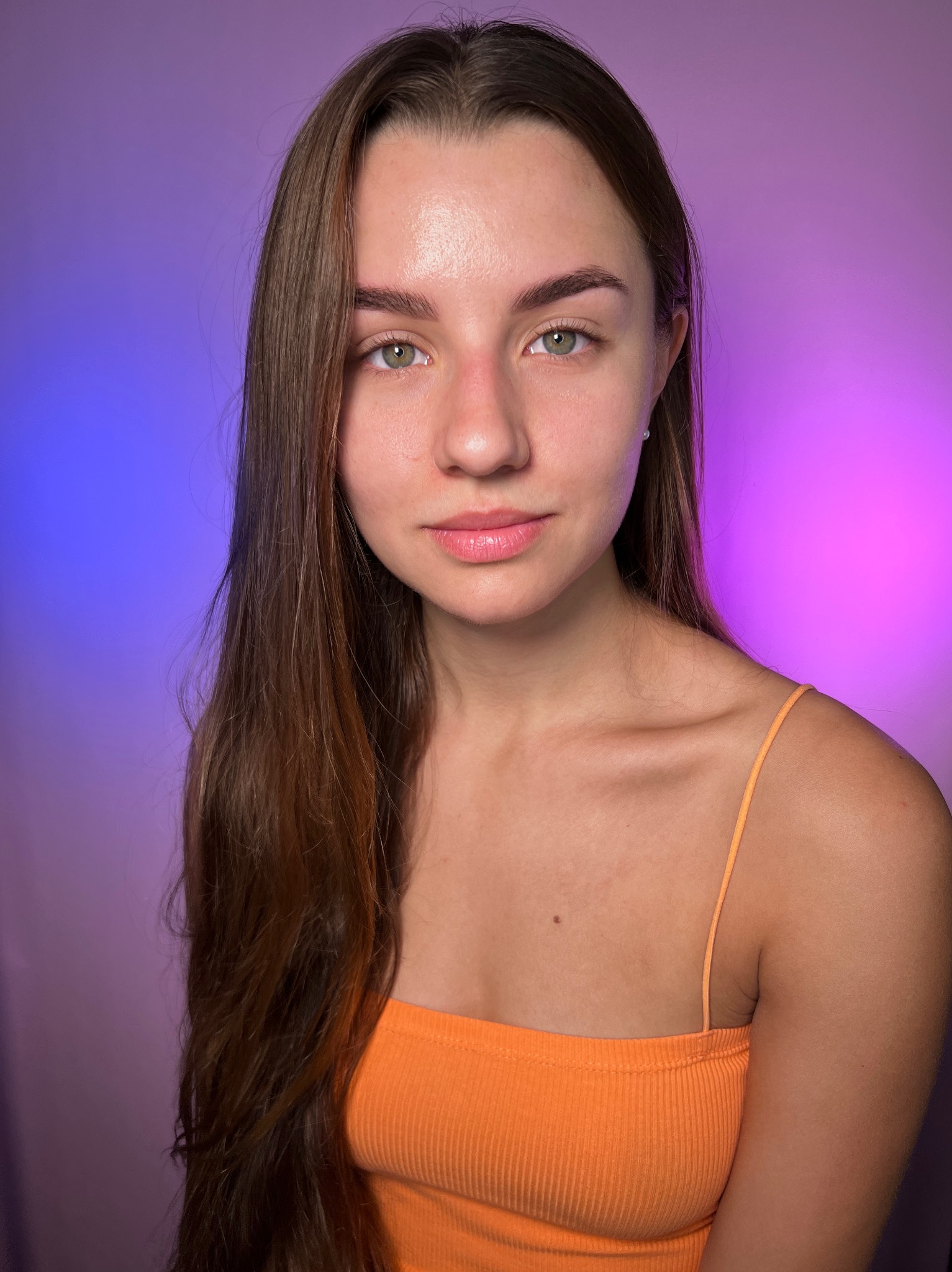 Portret kobiety z delikatnym makijażem, podkreślone brwi, naturalny odcień ust, długie brązowe włosy opadające na ramię, na tle fioletowo-różowego oświetlenia.