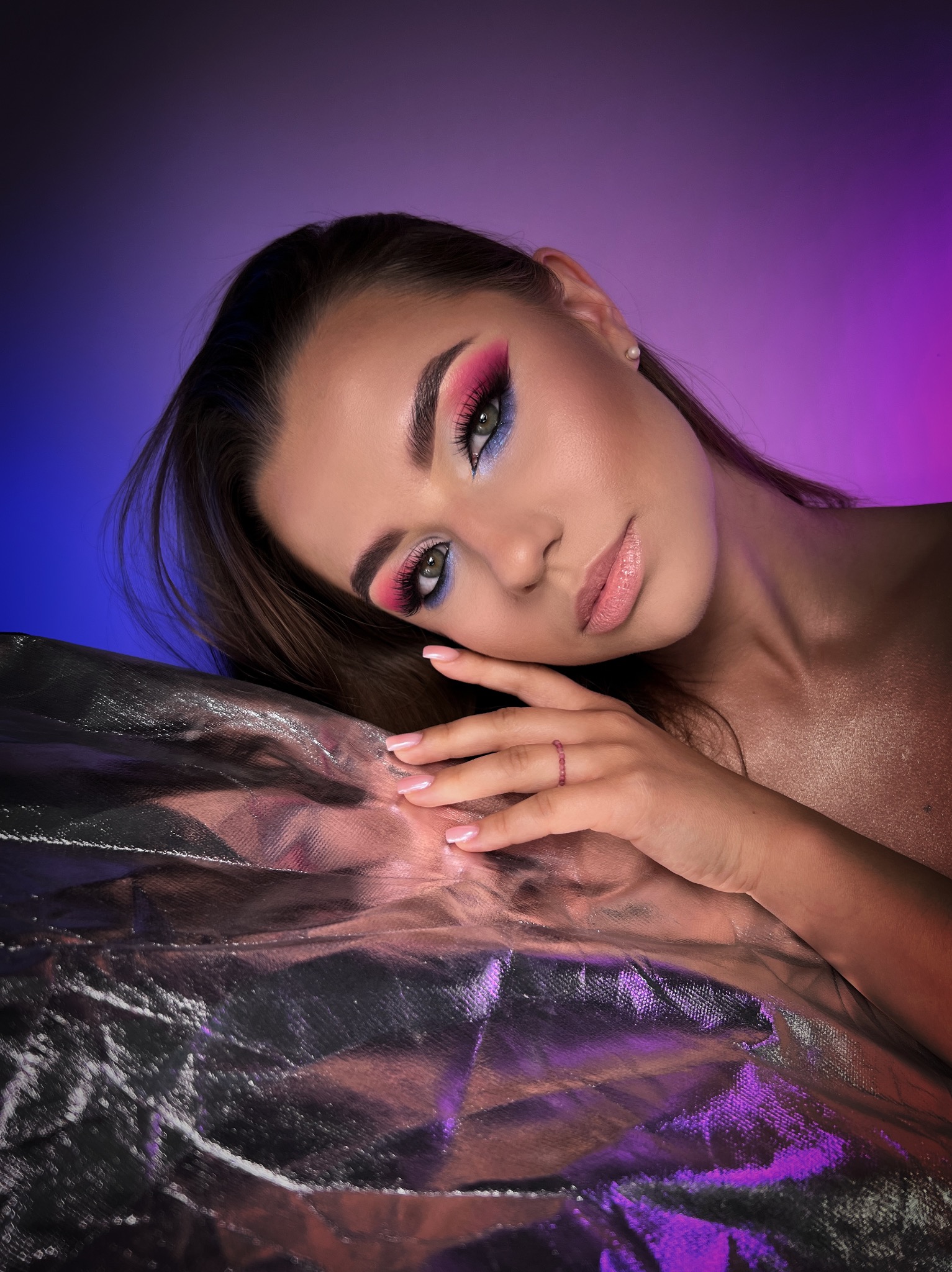 Portret kobiety z intensywnym, kolorowym wizażem oczu w odcieniach różu i błękitu, opierającej się o srebrzystą, połyskującą powierzchnię na tle gradientu fioletu i błękitu.
