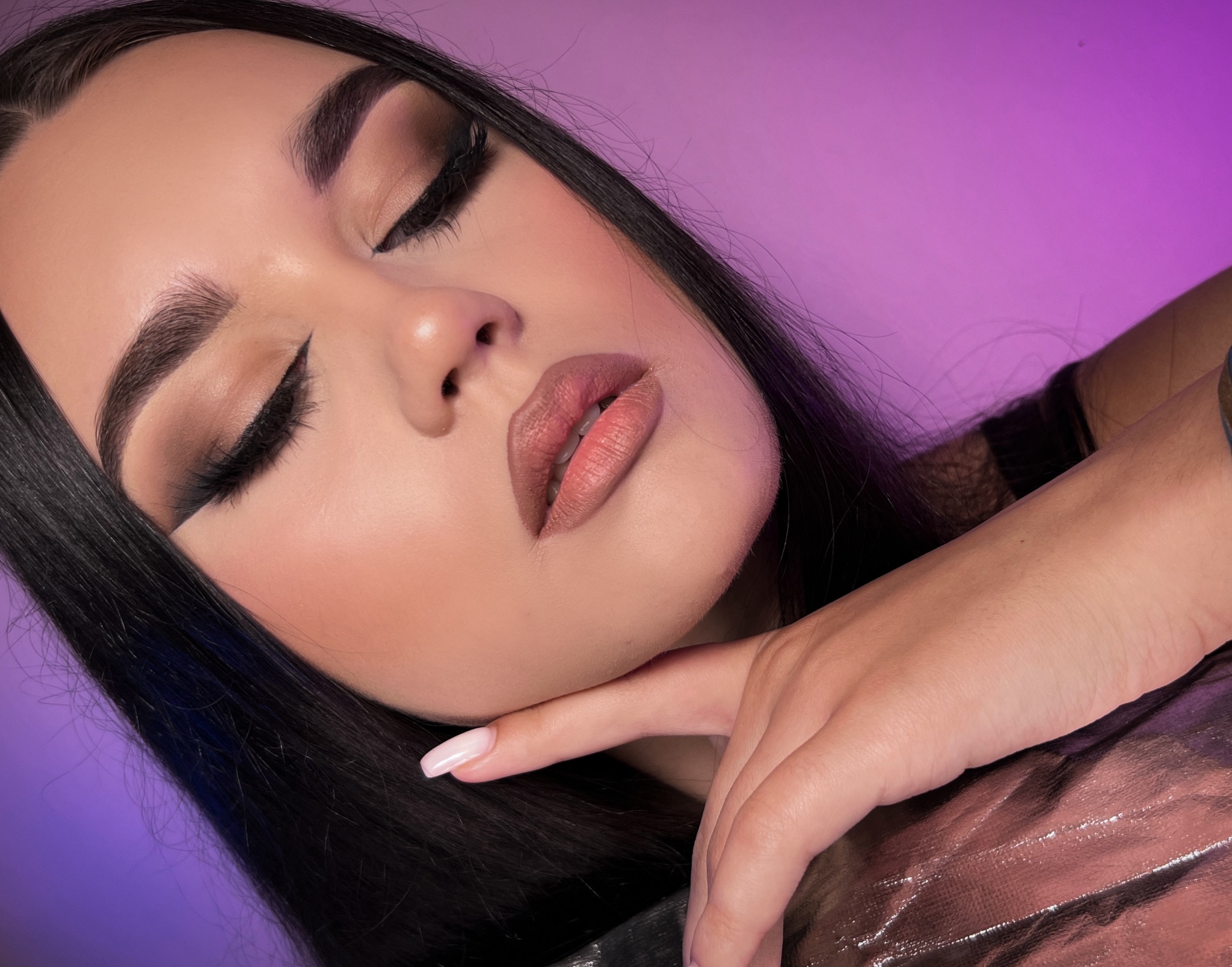 Portret kobiety z zamkniętymi oczami, prezentujący wyrazisty makijaż oczu w odcieniach brązu i idealnie podkreślone usta w kolorze nude, na tle w odcieniach fioletu.