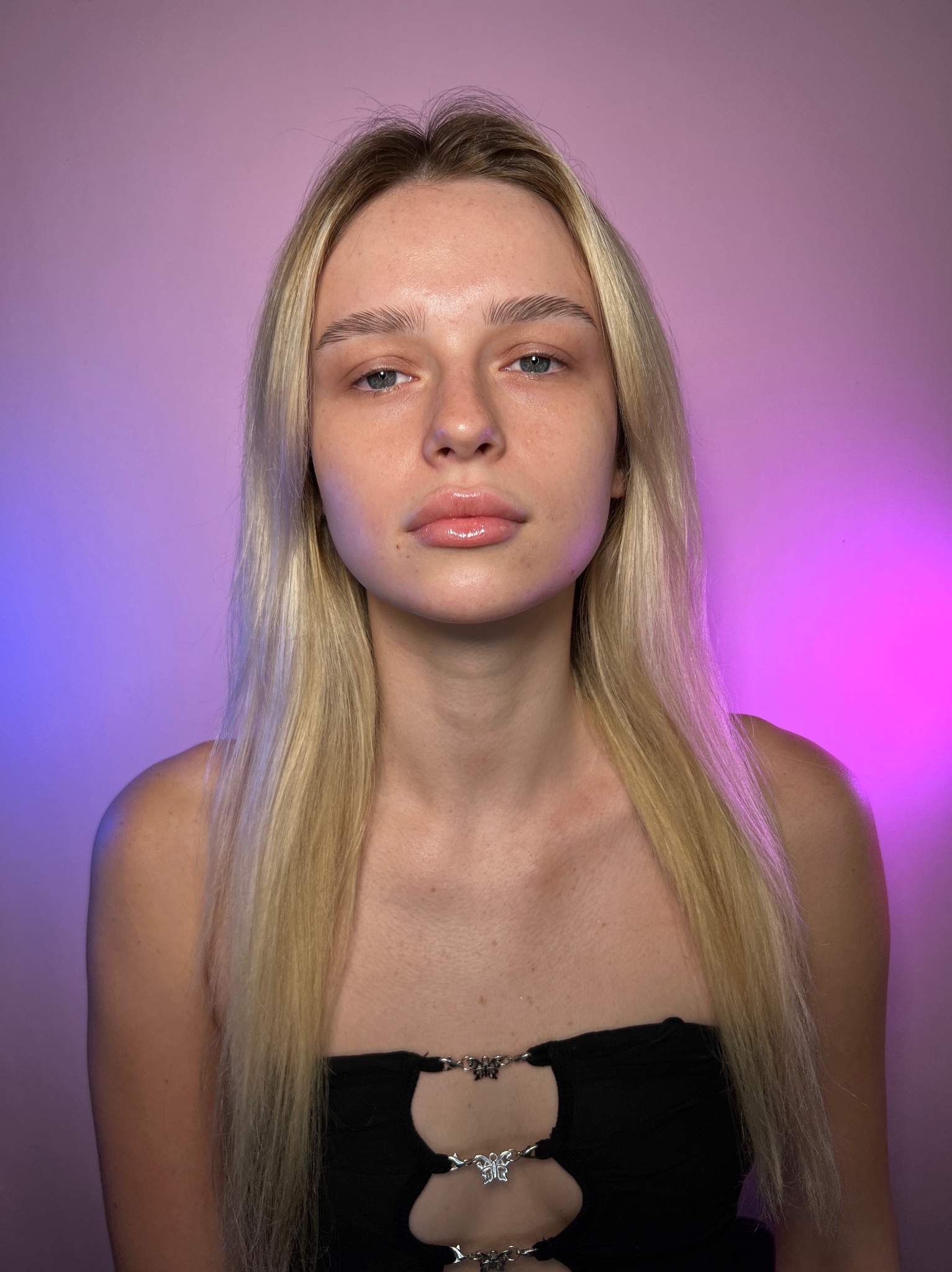Portret młodej kobiety z blond włosami i delikatnie błyszczącymi ustami na tle gradientu różowo-fioletowego.