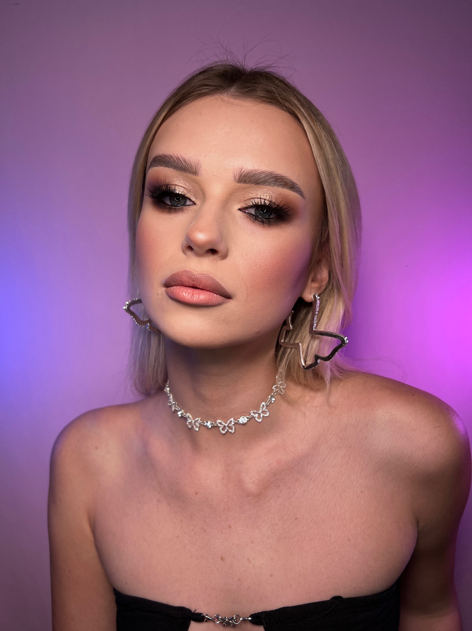 Portret blondynki z profesjonalnie wykonanym, błyszczącym makijażem oczu i ustami w odcieniu nude, srebrny naszyjnik i kolczyki w kształcie motyli, fioletowe tło.