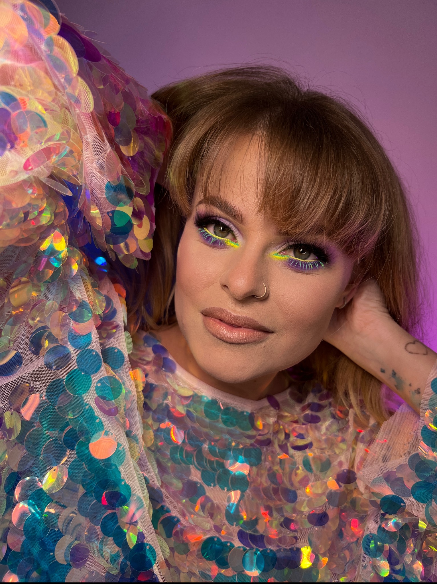 Portret kobiety z intensywnym makijażem oczu w odcieniach fioletu i neonowej zieleni, noszącej błyszczącą sukienkę z cekinami, na fioletowym tle.