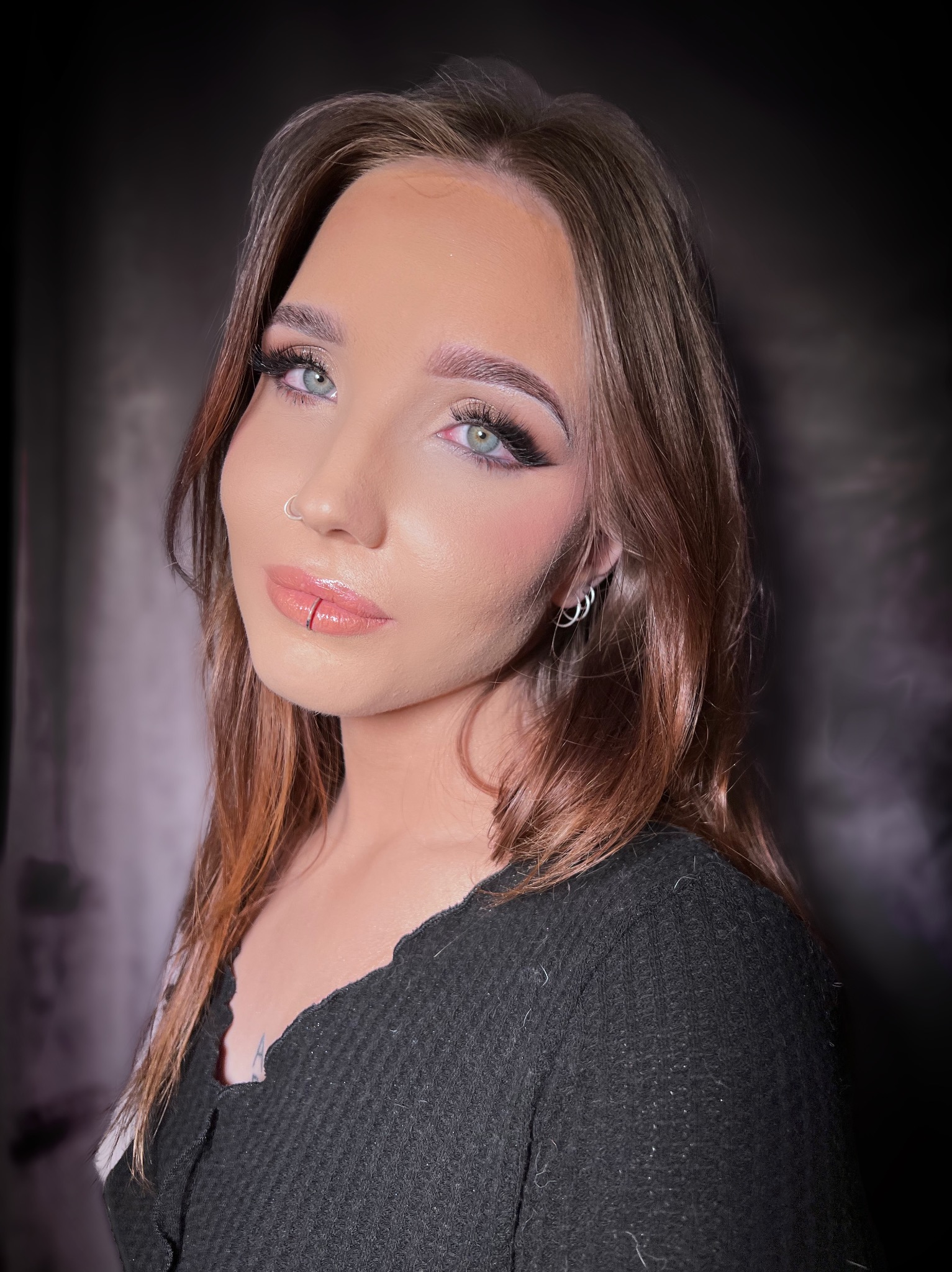 Portret kobiety z delikatnym konturowaniem, podkreślonymi oczami z czarnym eyelinerem i błyszczącymi ustami z kolczykiem vertical labret.