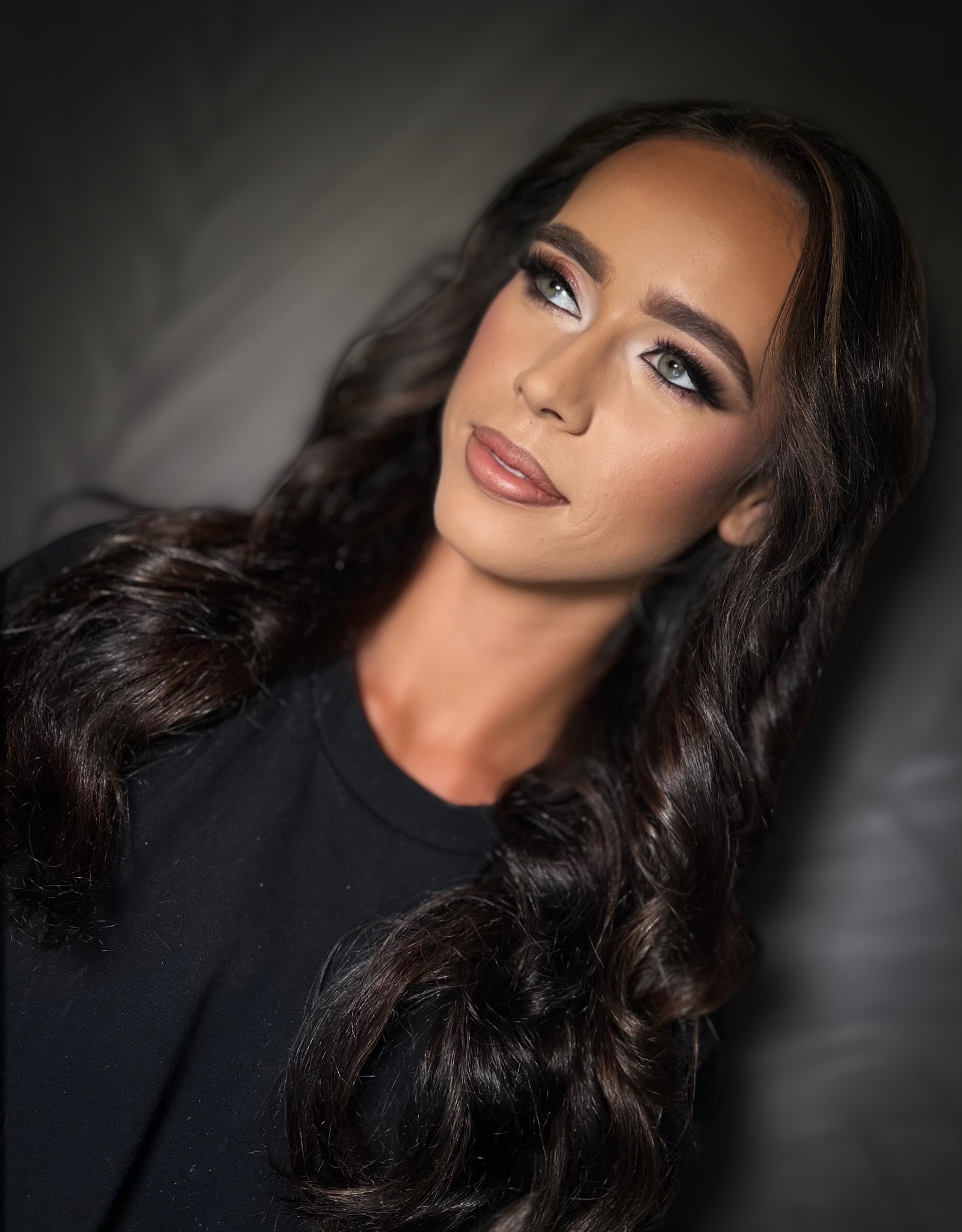 Portret kobiety z profesjonalnie wykonanym konturowaniem twarzy i subtelnym podkreśleniem oczu, spoglądającej w górę, z ciemnymi, falowanymi włosami na ciemnym tle.
