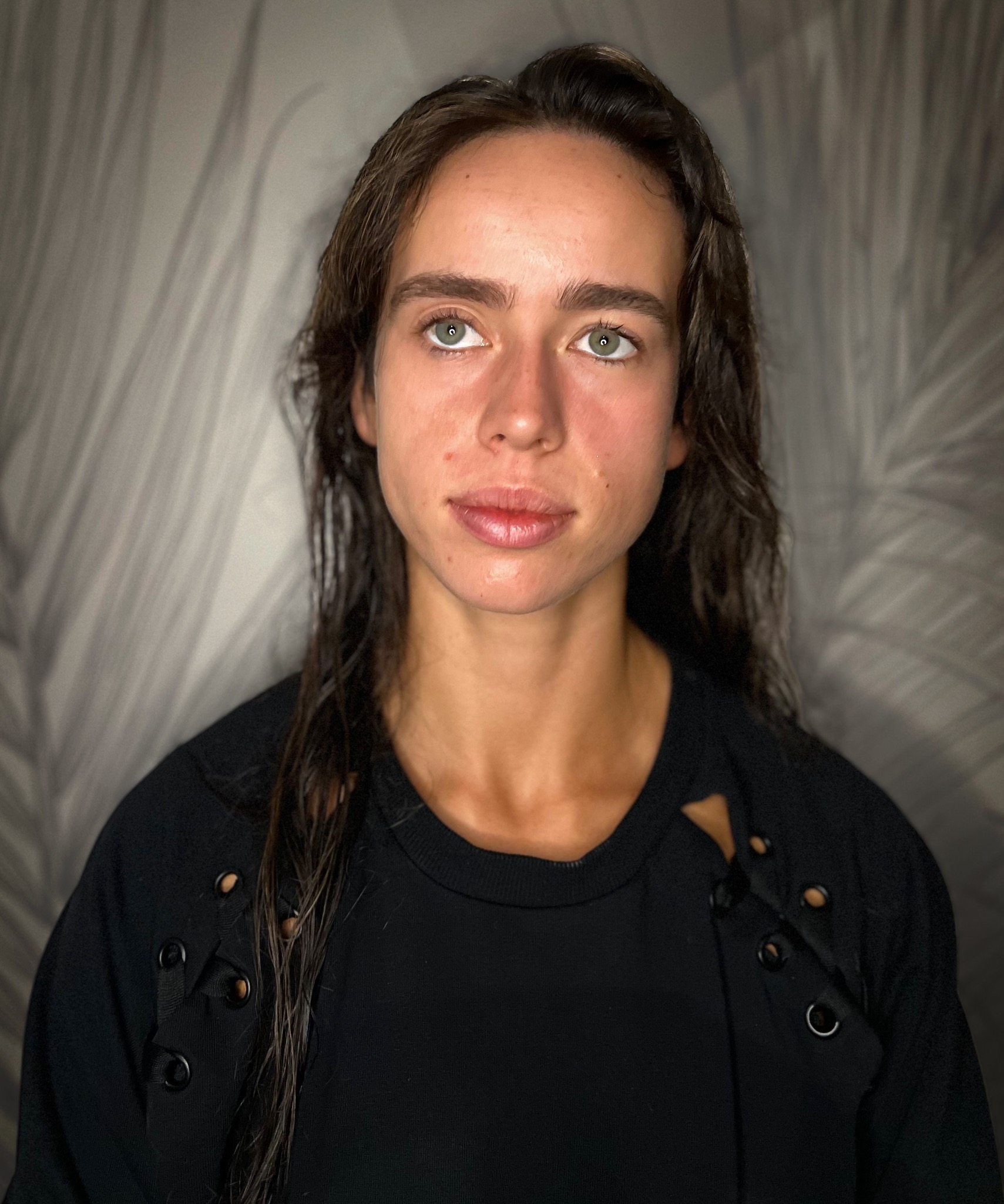 Portret kobiety przed nałożeniem makijażu, z mokrymi włosami i widocznymi niedoskonałościami skóry, spoglądającej w górę.