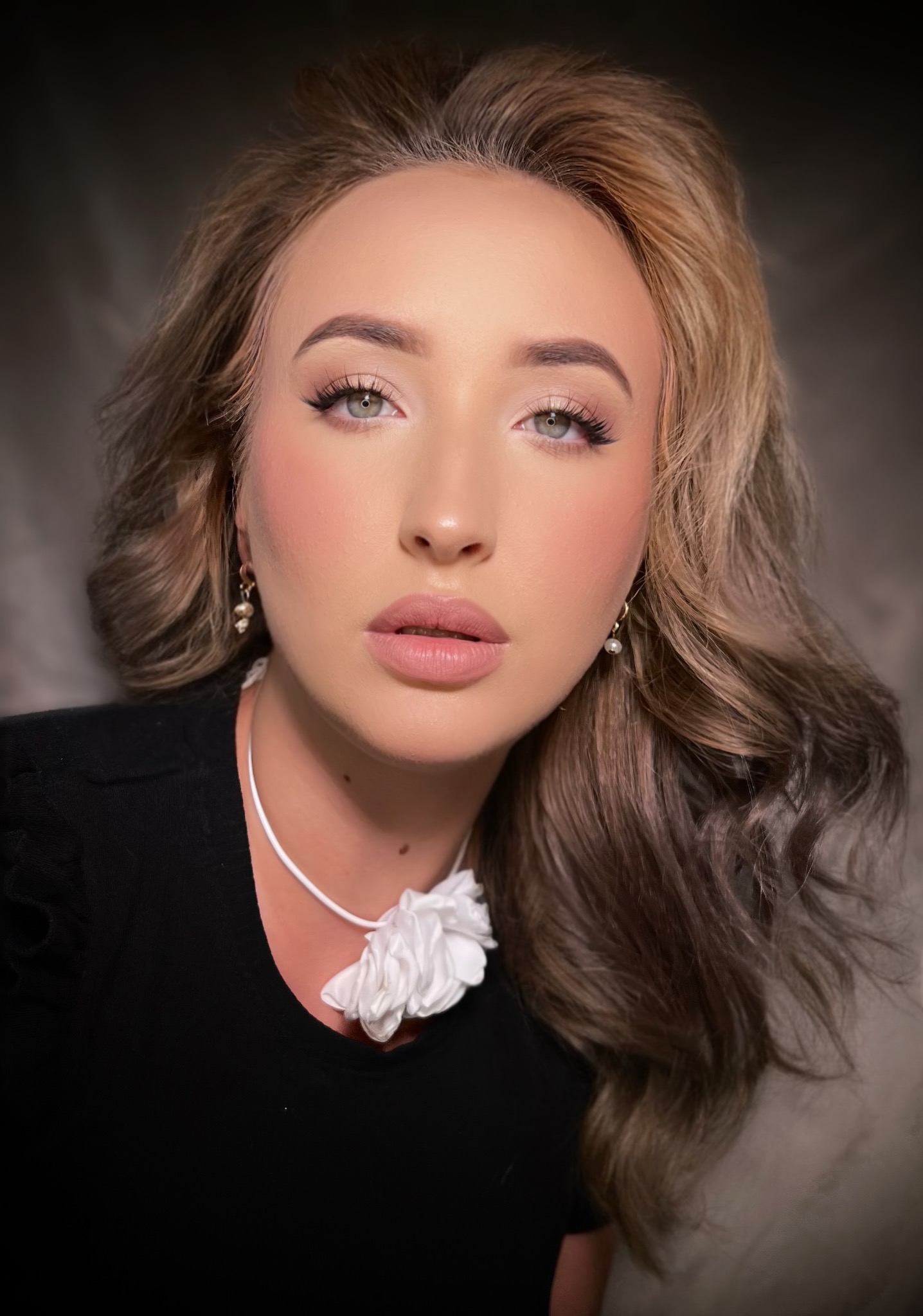 Portret kobiety z delikatnym, profesjonalnym upiększeniem, podkreślonymi rzęsami i ustami w odcieniu nude, uzupełnionym przez perłowe kolczyki i biały kwiat przy szyi.