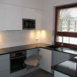 Nowoczesna kuchnia z białymi szafkami, czarnym blatem i narożną półką obrotową, widok na budynek przez okno z żaluzjami.